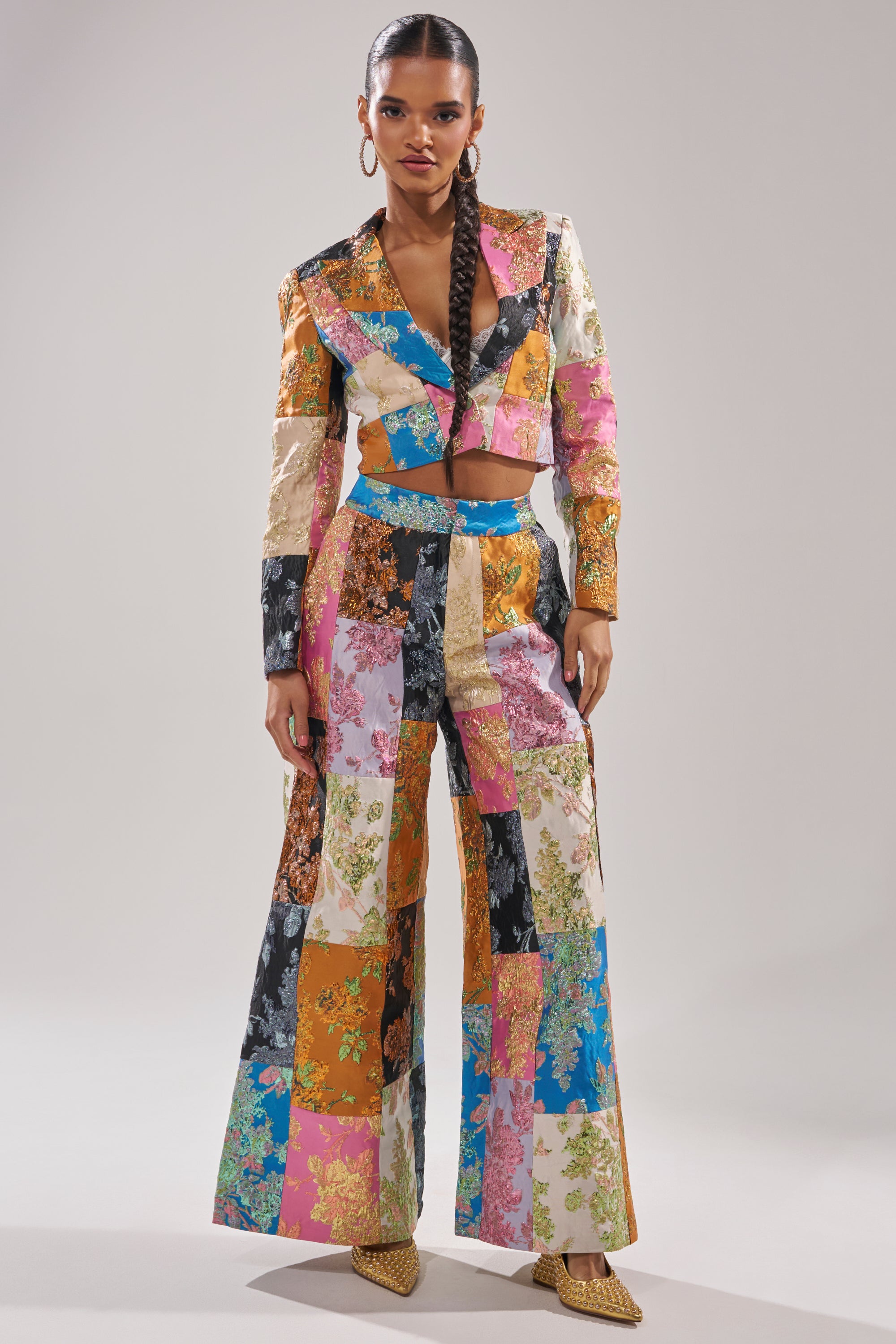 A woman models the LOLITA PATCHWORK BROCADE TROUSER, featuring colorful patchwork and floral patterns with wide legs. She pairs it with a cropped blazer, gold earrings, and long braids against a neutral backdrop. 0
