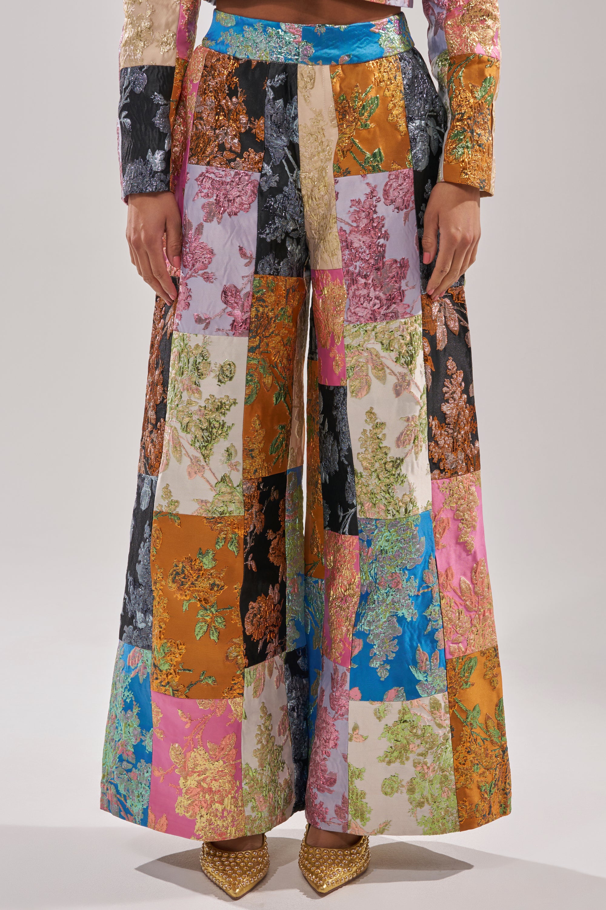 Someone in LOLITA PATCHWORK BROCADE TROUSER, featuring a vibrant floral brocade design, pairs them with gold pointed-toe shoes and stands before a simple light backdrop. 1
