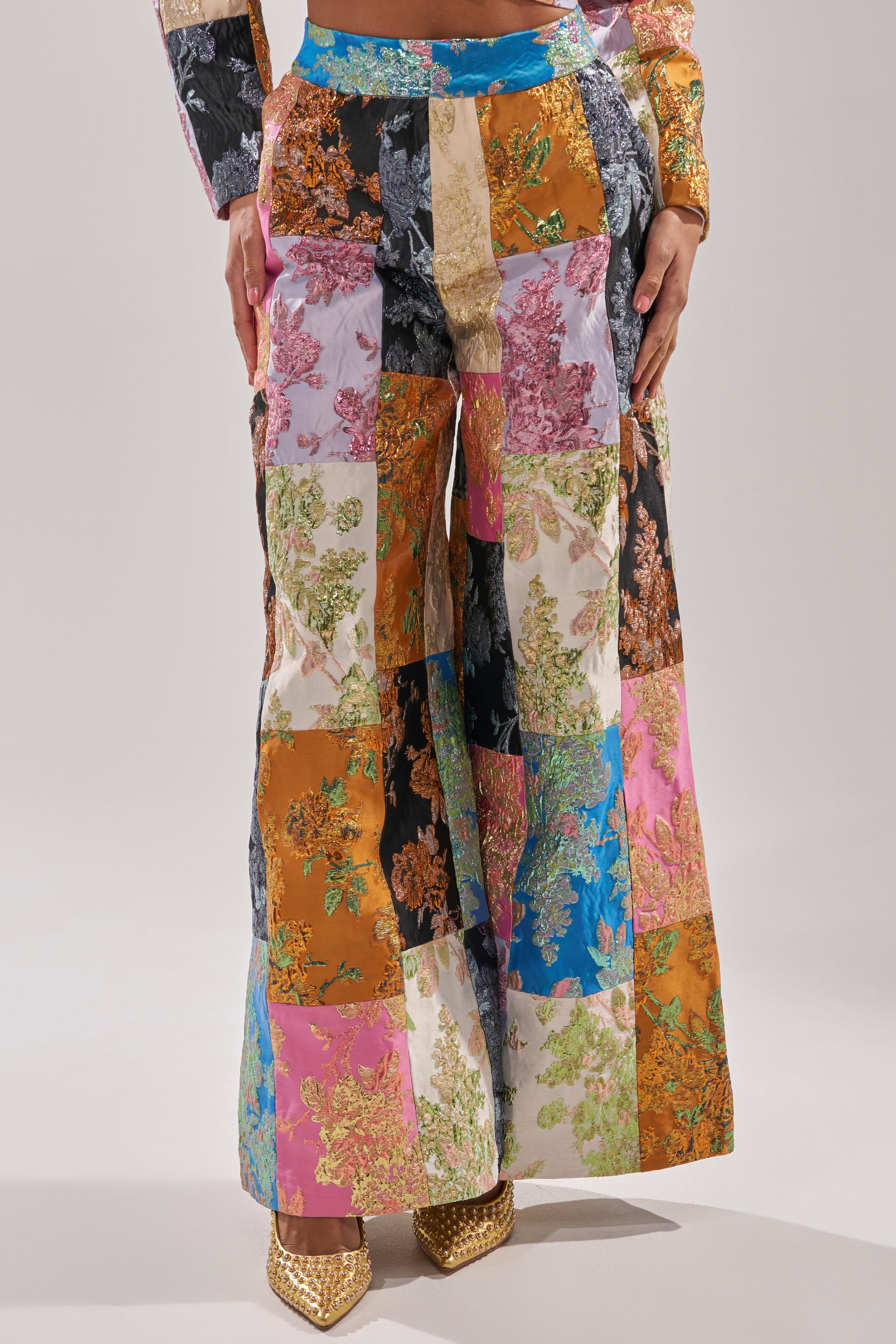 A person models the LOLITA PATCHWORK BROCADE TROUSER, featuring colorful patchwork and gold floral designs, paired with gold pointed-toe heels. A matching sleeve and hand are also visible against a plain background. 2