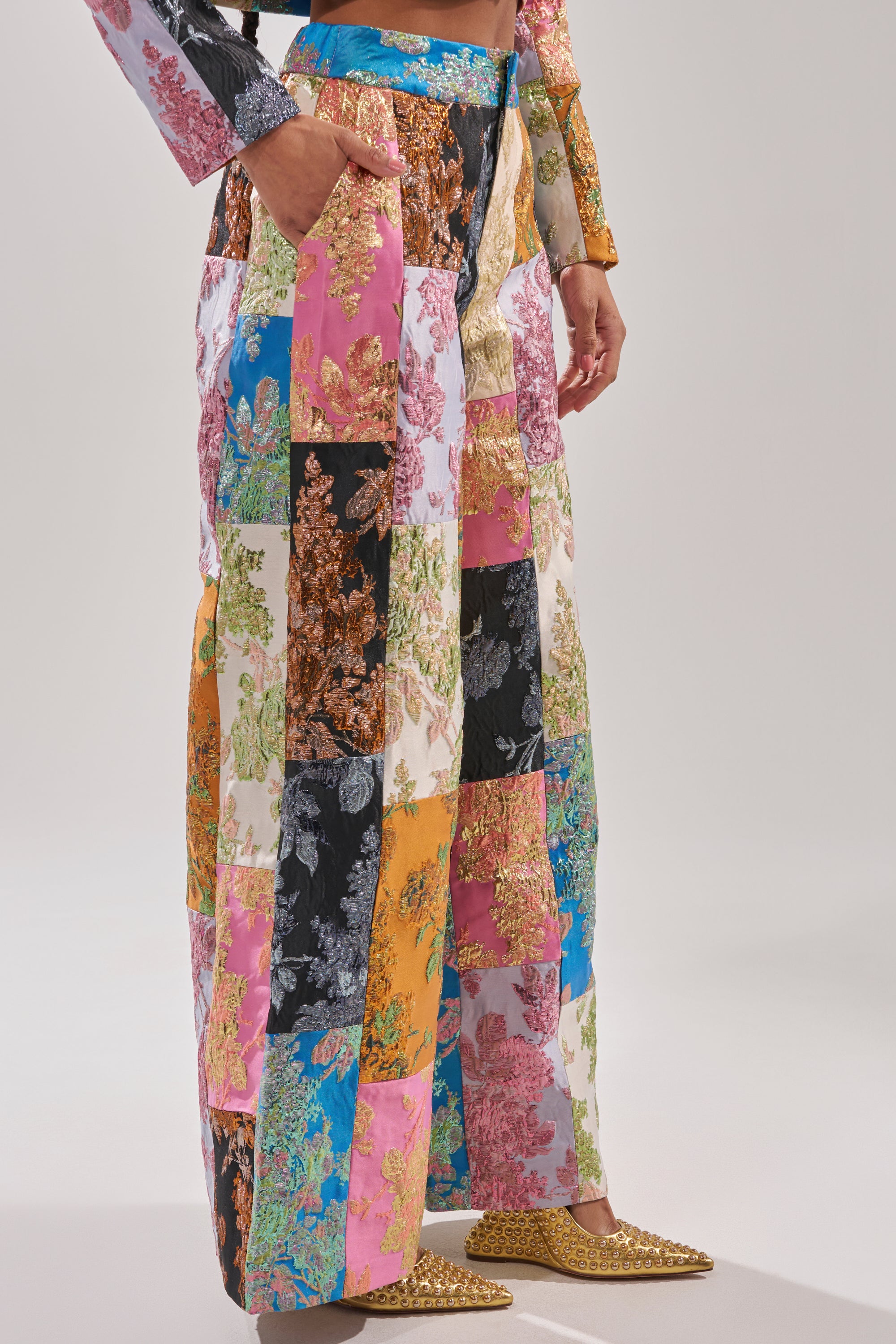Wearing the LOLITA PATCHWORK BROCADE TROUSER styled as wide-leg pants with a matching top and gold studded flats, a person stands against a plain background. 3