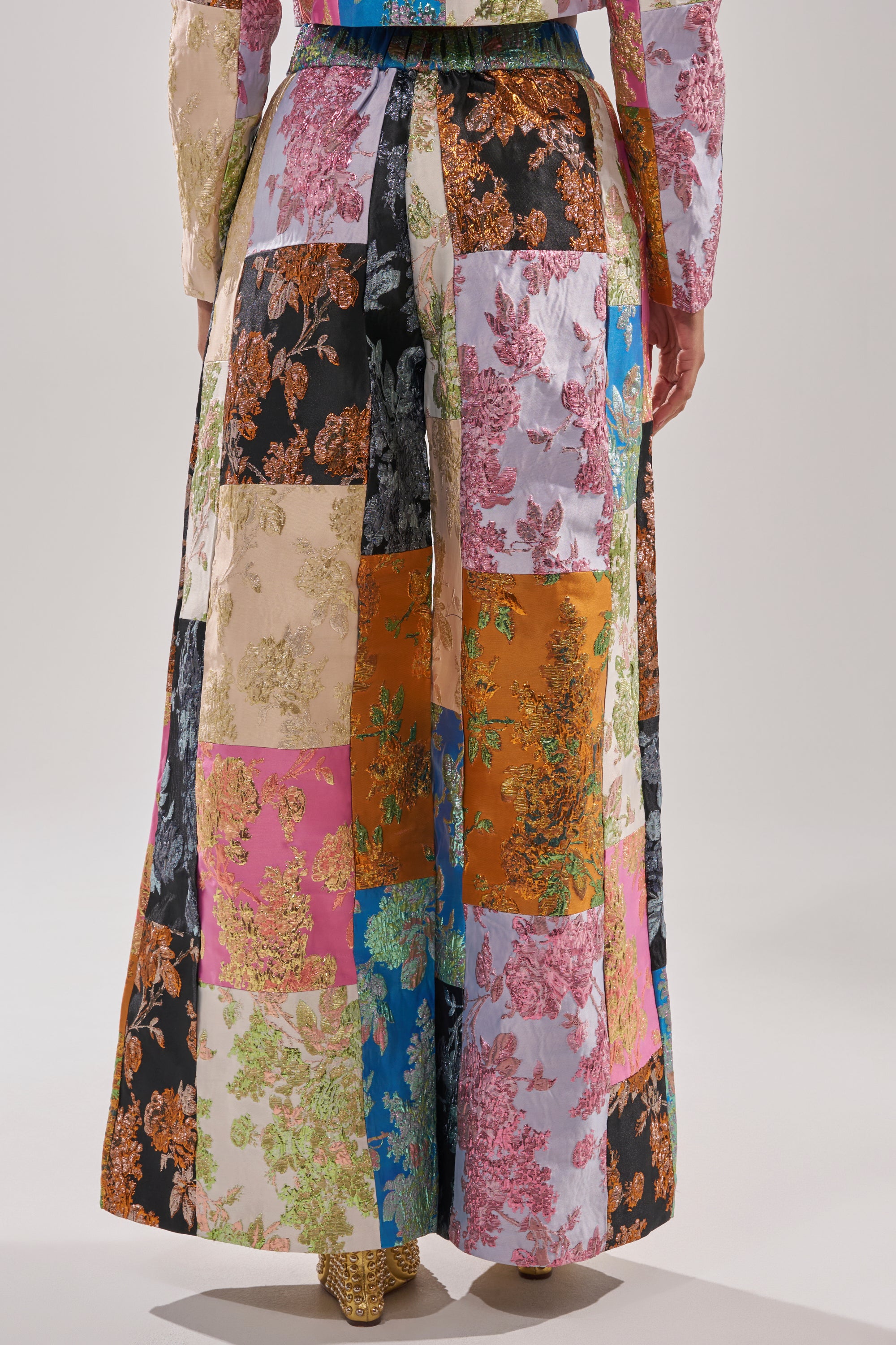 A person wearing a long skirt of colorful floral patchwork, inspired by the LOLITA PATCHWORK BROCADE TROUSER, is photographed from the back against a plain background. 4