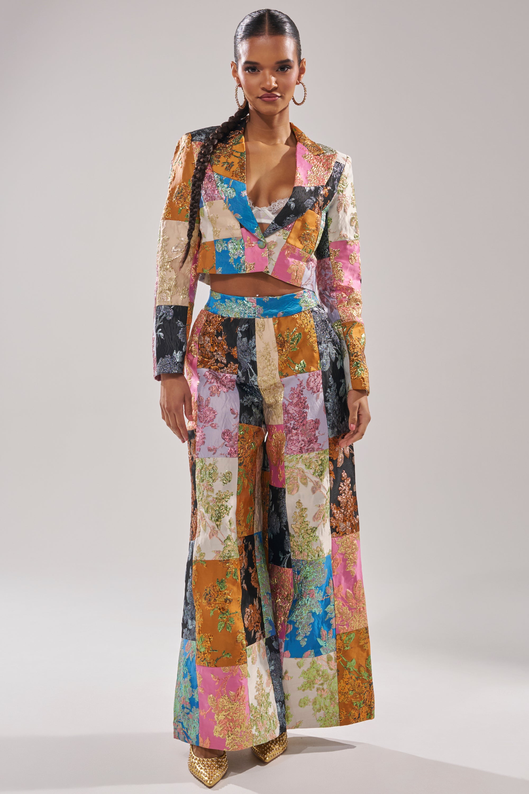 A woman stands facing forward, wearing a vibrant patchwork suit with a cropped blazer and the LOLITA PATCHWORK BROCADE TROUSER in colorful floral squares. She has long braids, hoop earrings, and gold pointed-toe shoes. 5