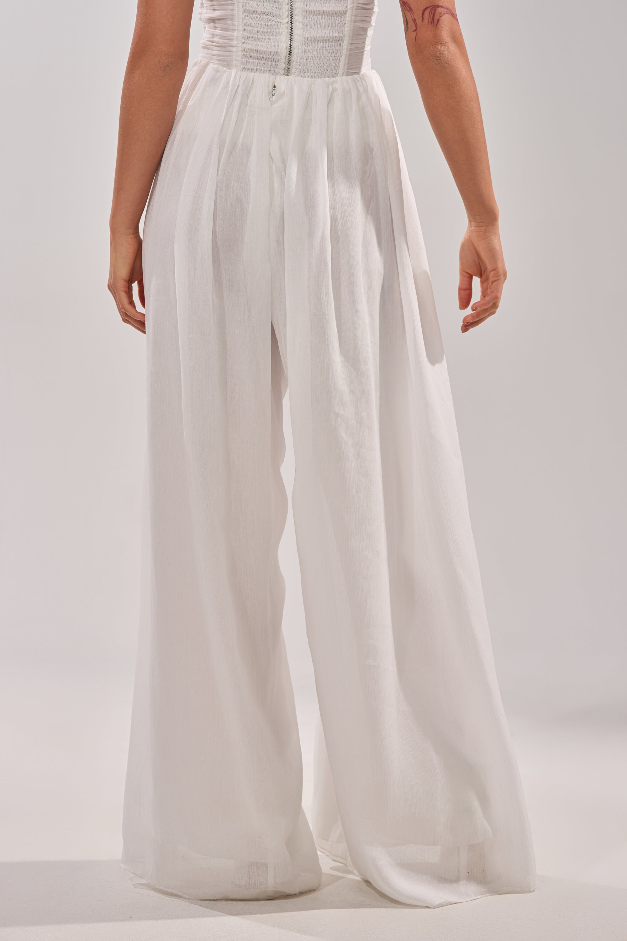 A person stands with their back to the camera, wearing the BEAUTIFUL WHITE PALAZZO PANT—high-waisted and sheer with flowing wide legs—paired with a sleeveless white top, against a plain light background. 3