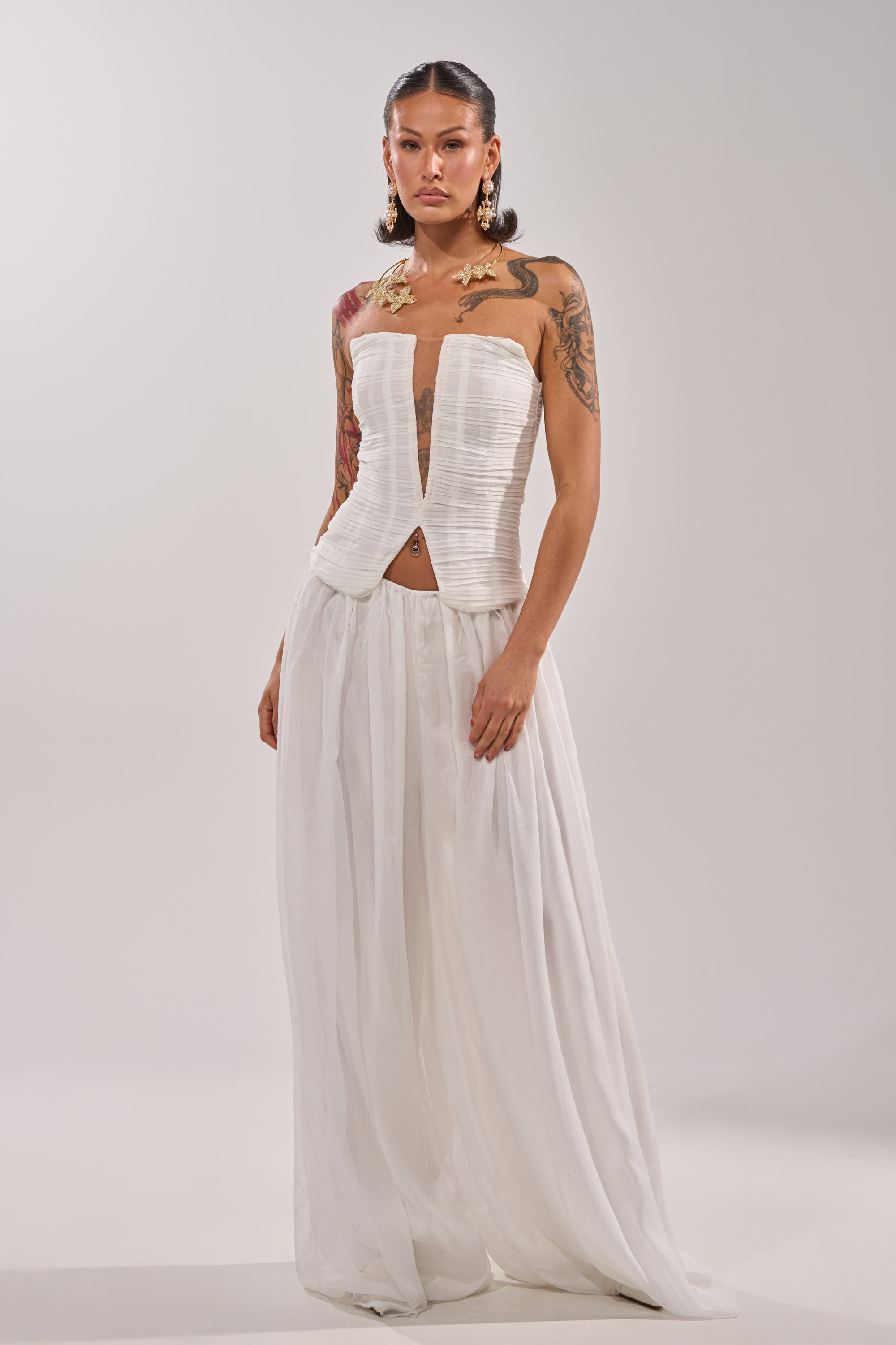 A woman with tattoos wears the BEAUTIFUL WHITE PALAZZO PANT, featuring a strapless fitted bodice and sheer, wide-leg skirt. Gold butterfly accessories decorate her shoulders, and she pairs it with large earrings against a plain light background. 5
