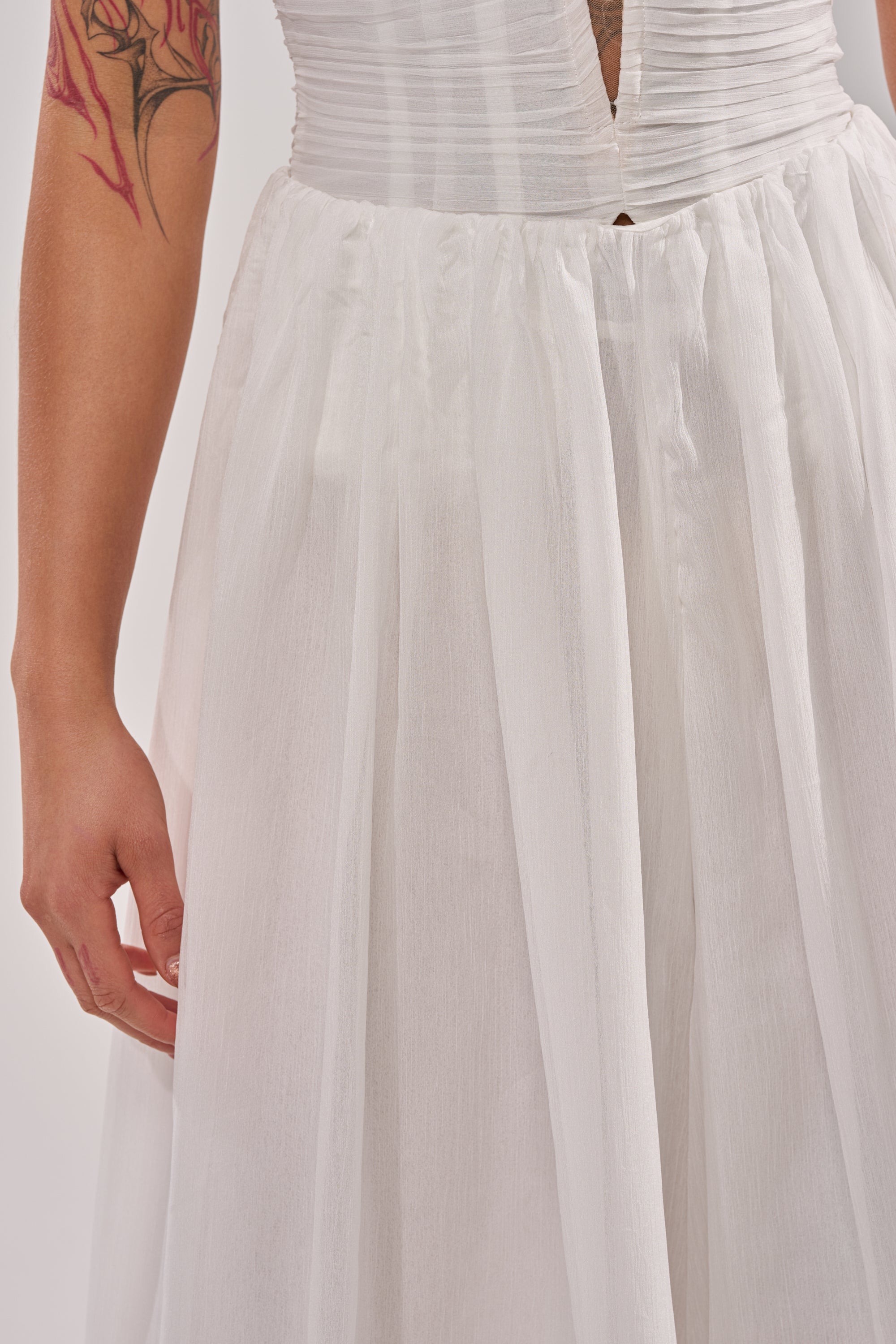 A close-up of a person wearing the BEAUTIFUL WHITE PALAZZO PANT, styled with a white sleeveless, pleated deep V-neck top. The tattooed arm and relaxed pose highlight an effortless look, ideal for wide-leg silhouettes and easy elegance. 6