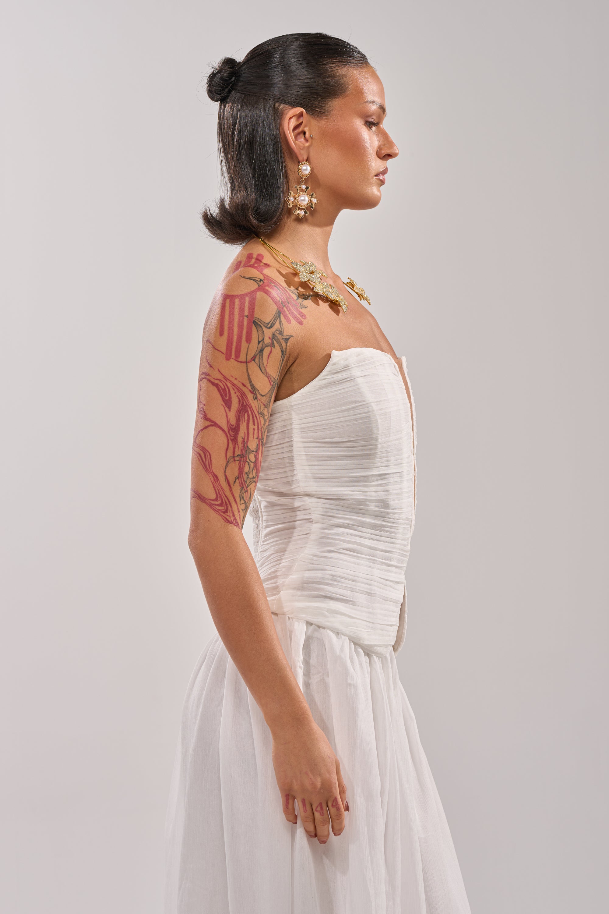 A woman with dark hair pulled back and a tattoo sleeve stands in profile against a light background, wearing the BEAUTIFUL NIGHT WHITE CORSET, paired with large earrings and a gold necklace. 2