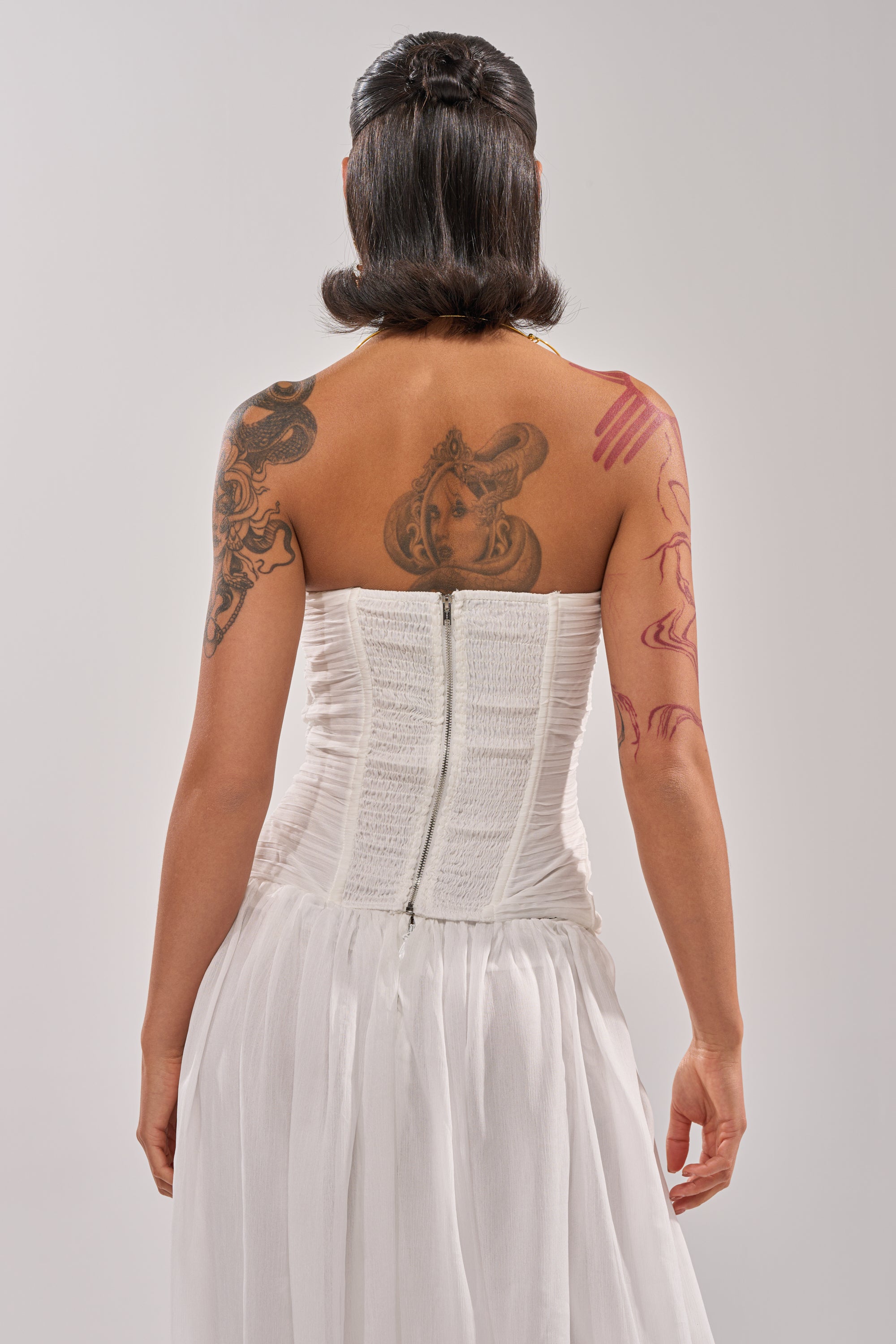 A woman with short dark hair and visible tattoos on her back and arm is seen from behind, wearing the BEAUTIFUL NIGHT WHITE CORSET—a strapless dress featuring a zippered back and flowing skirt. 3