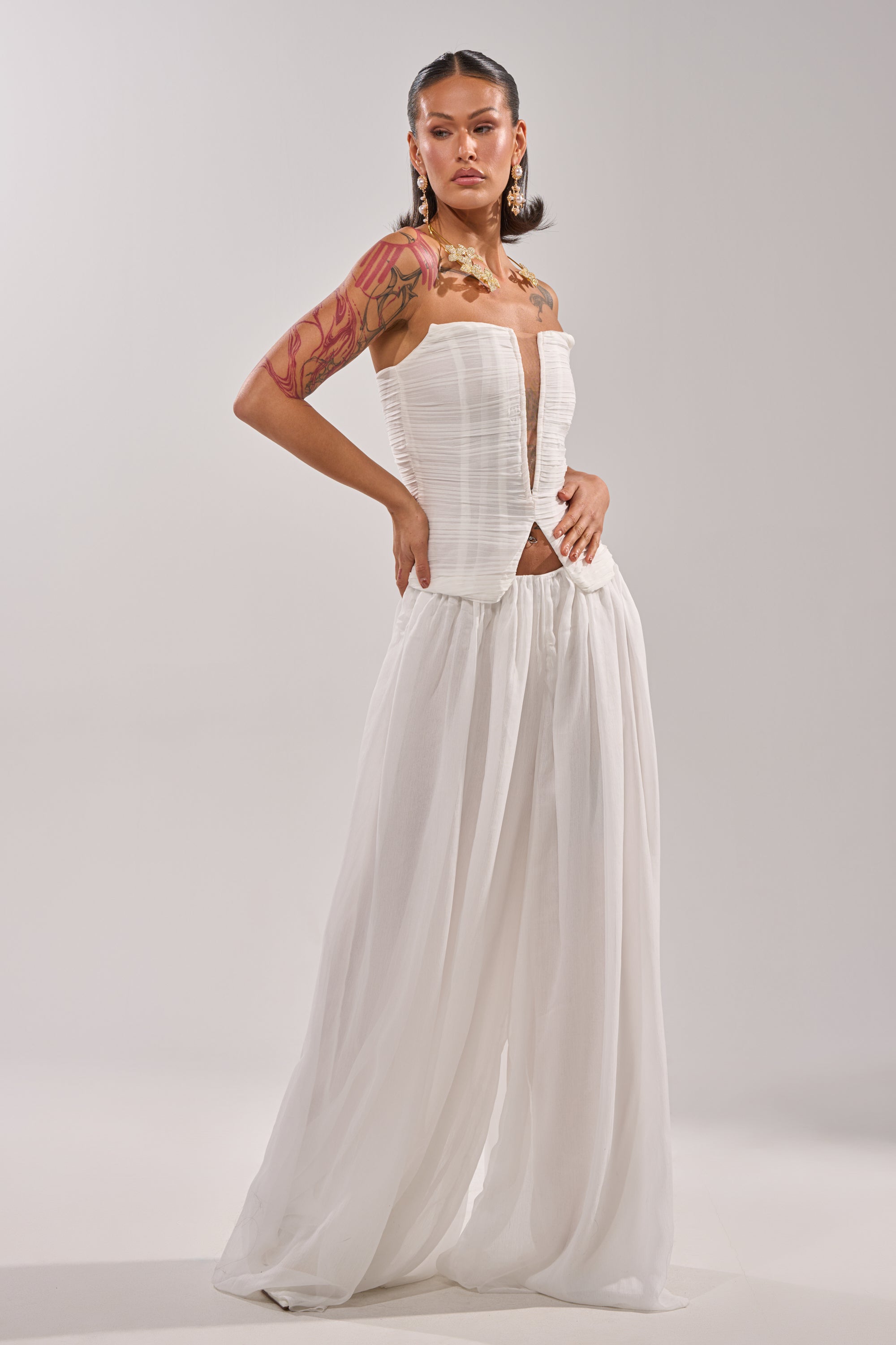 A woman poses confidently in the BEAUTIFUL NIGHT WHITE CORSET, a strapless white top with a structured bodice, paired with flowy wide-leg white pants. She showcases arm tattoos and large earrings against a plain light background. 4