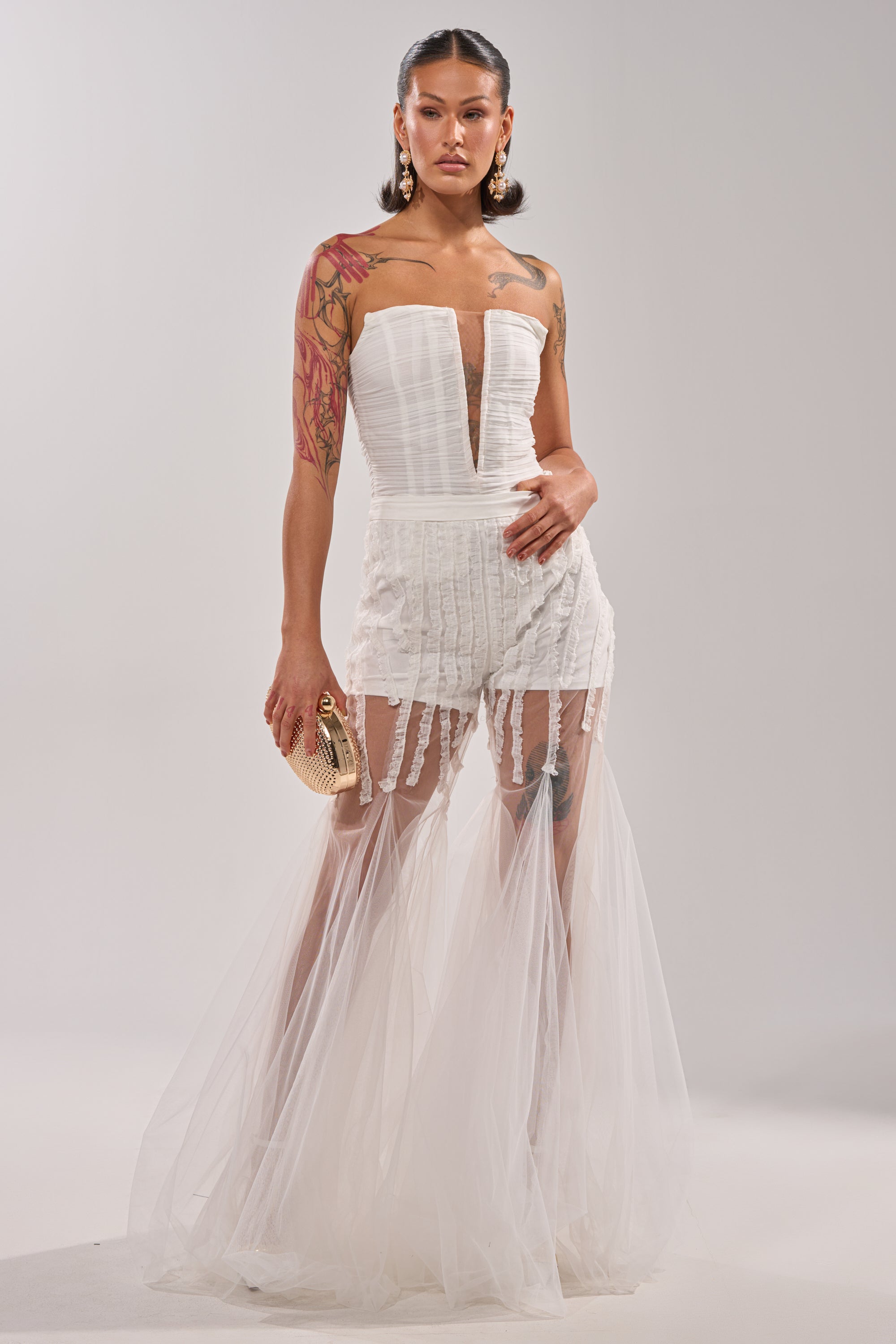 She wears the BEAUTIFUL NIGHT WHITE CORSET, featuring a strapless design, deep neckline, sheer mesh skirt, and lace shorts. Her look includes visible tattoos, large earrings, a small gold clutch, and slicked-back hair against a plain background. 6