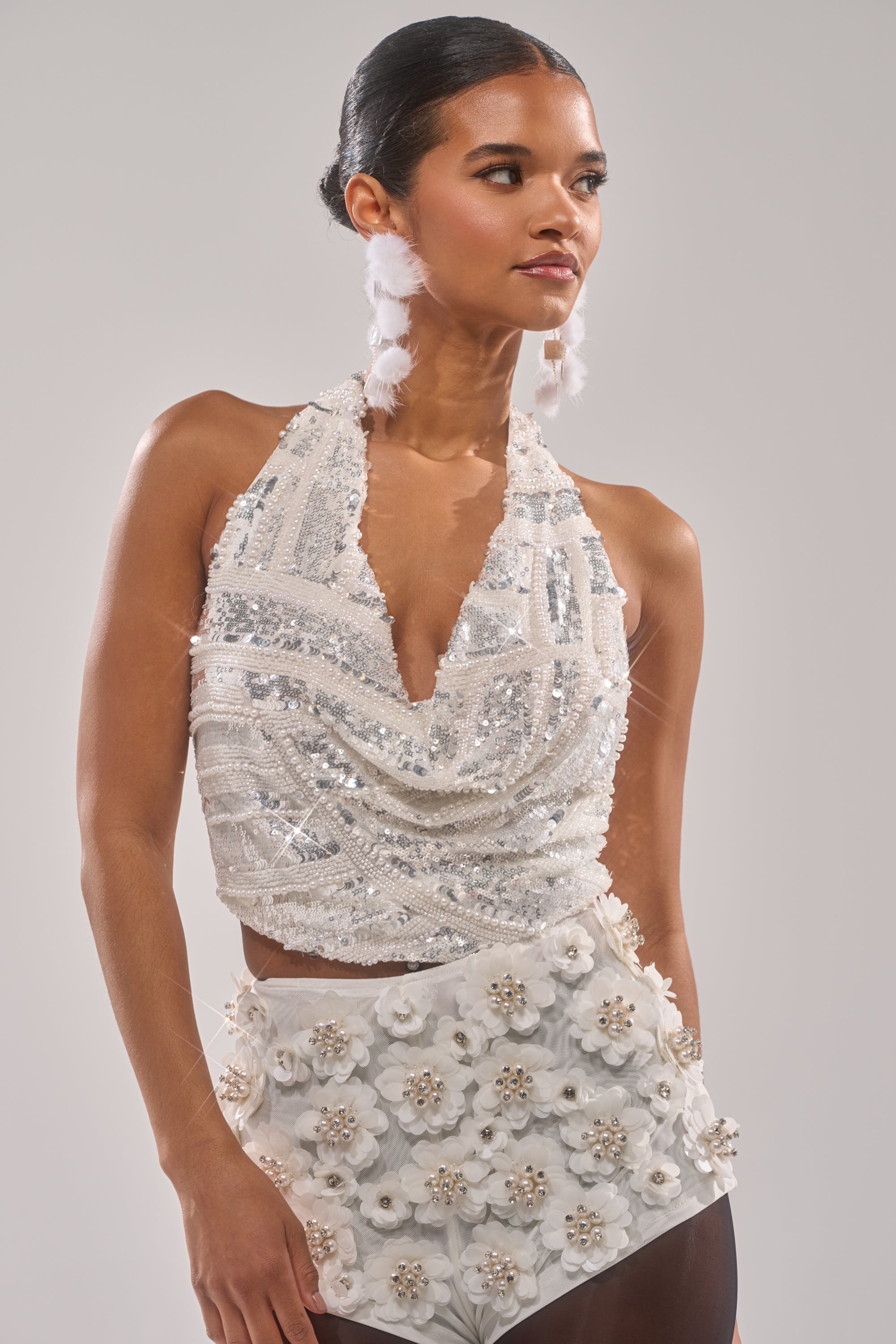 A woman wearing the SHINE BRIGHT SEQUIN BLOUSE—pearl-embroidered, sleeveless, with a draped neckline—paired with a white mini skirt featuring 3D floral embellishments, stands against a plain background and looks to the side. 0