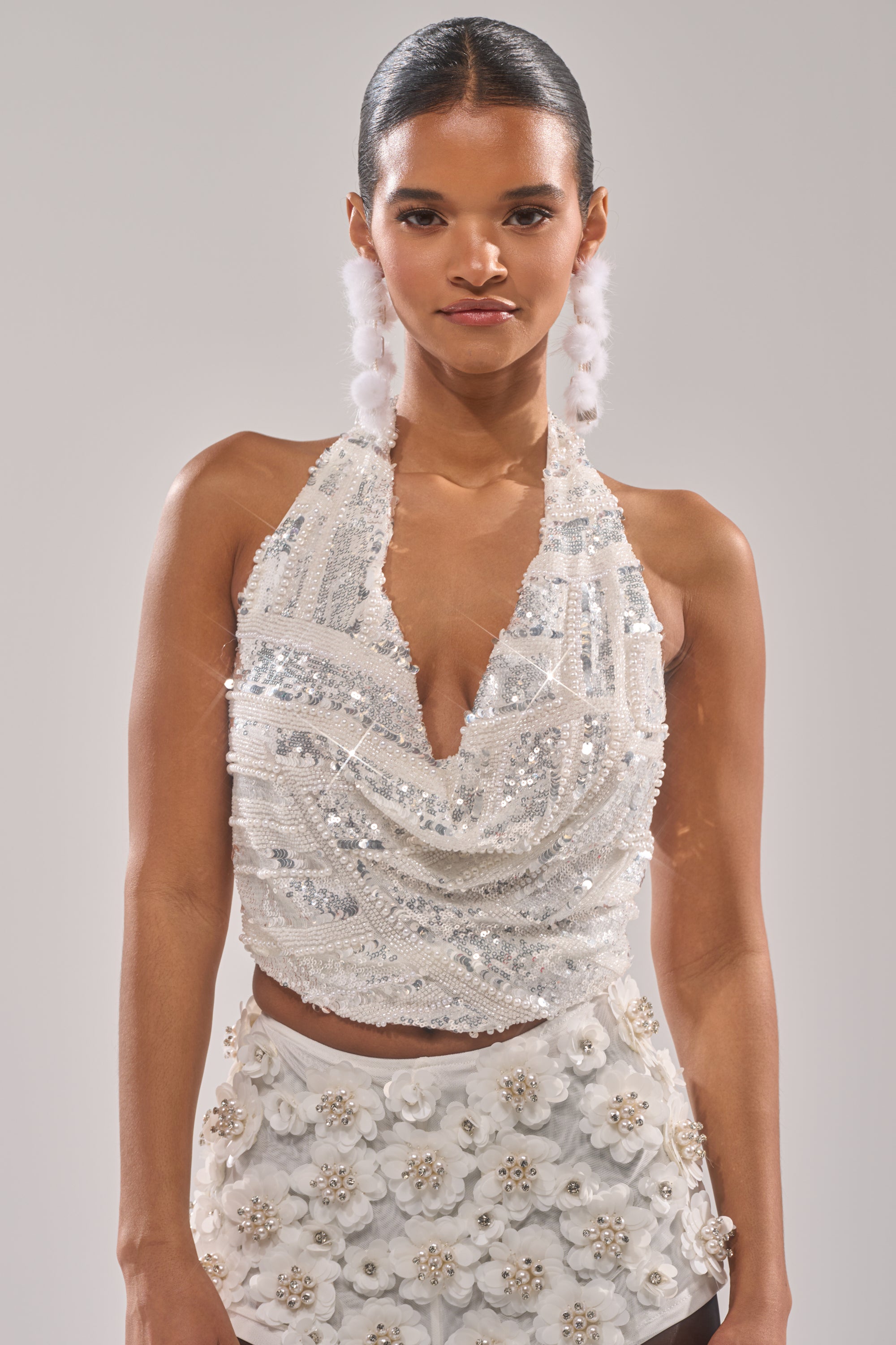 Wearing the SHINE BRIGHT SEQUIN BLOUSE with a halter neckline and a white 3D floral appliqué skirt, she completes her special occasion look with sleek hair and bold white pom-pom earrings. 2