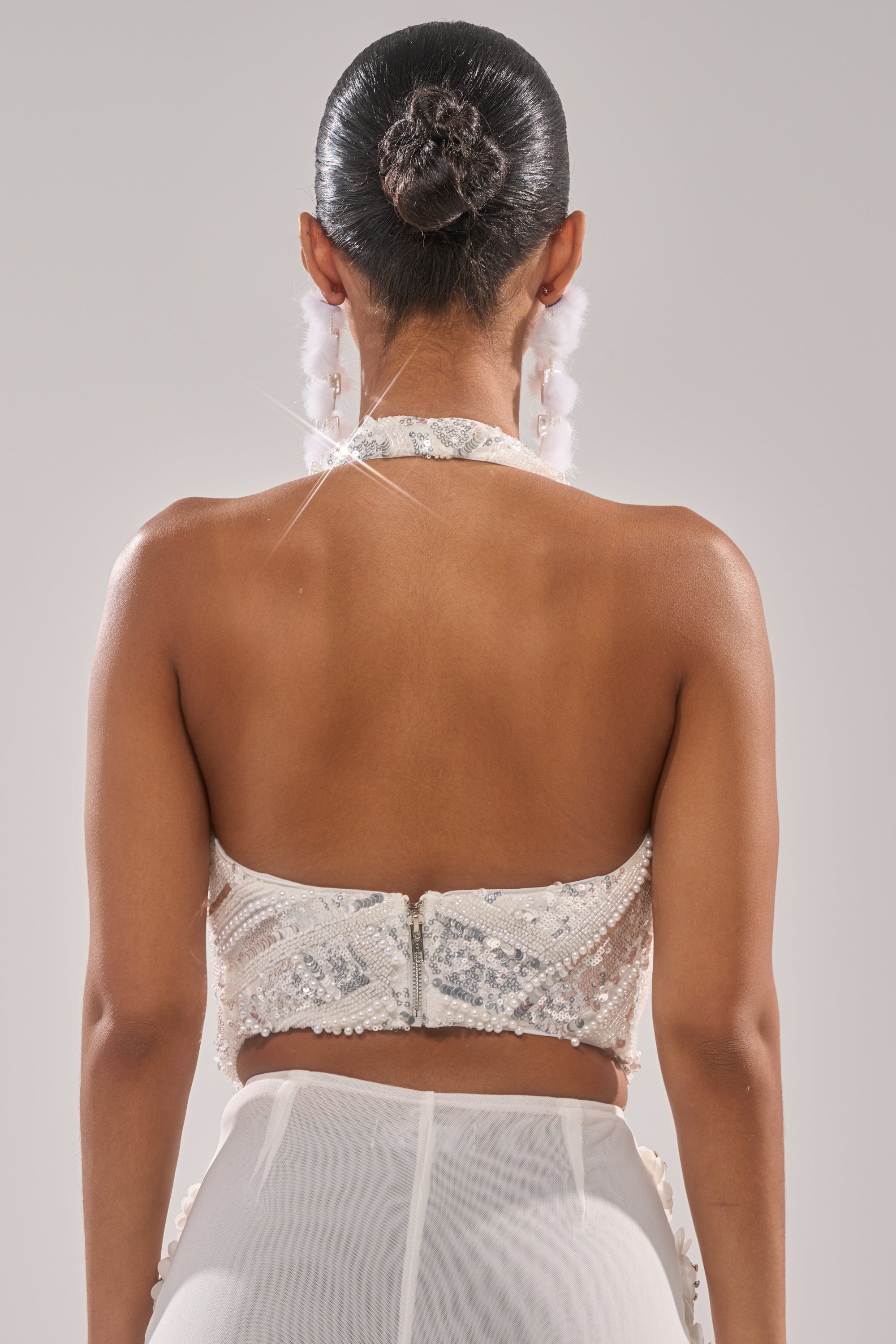 A woman with her hair in a bun is seen from behind wearing the SHINE BRIGHT SEQUIN BLOUSE, a cropped white sequin top, paired with sheer white pants and large white earrings, against a plain light gray background. 4