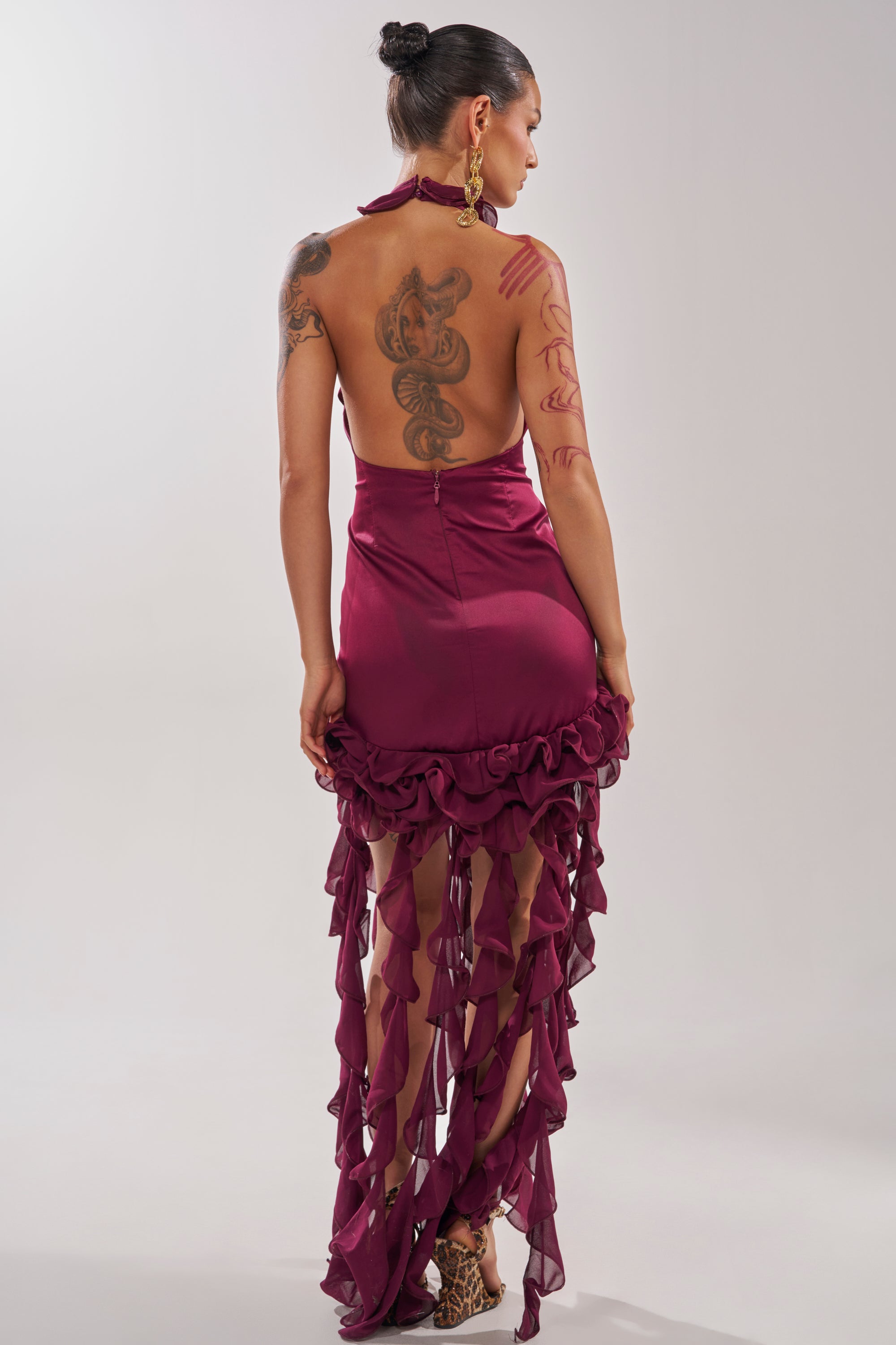 A woman faces away in the JET 2 HOLIDAY MINI DRESS—a burgundy halter with ruffle fringe and sheer, cascading fabric. Tattoos cover her arms and back, large gold earrings shine, and her hair is styled in a neat bun. 6