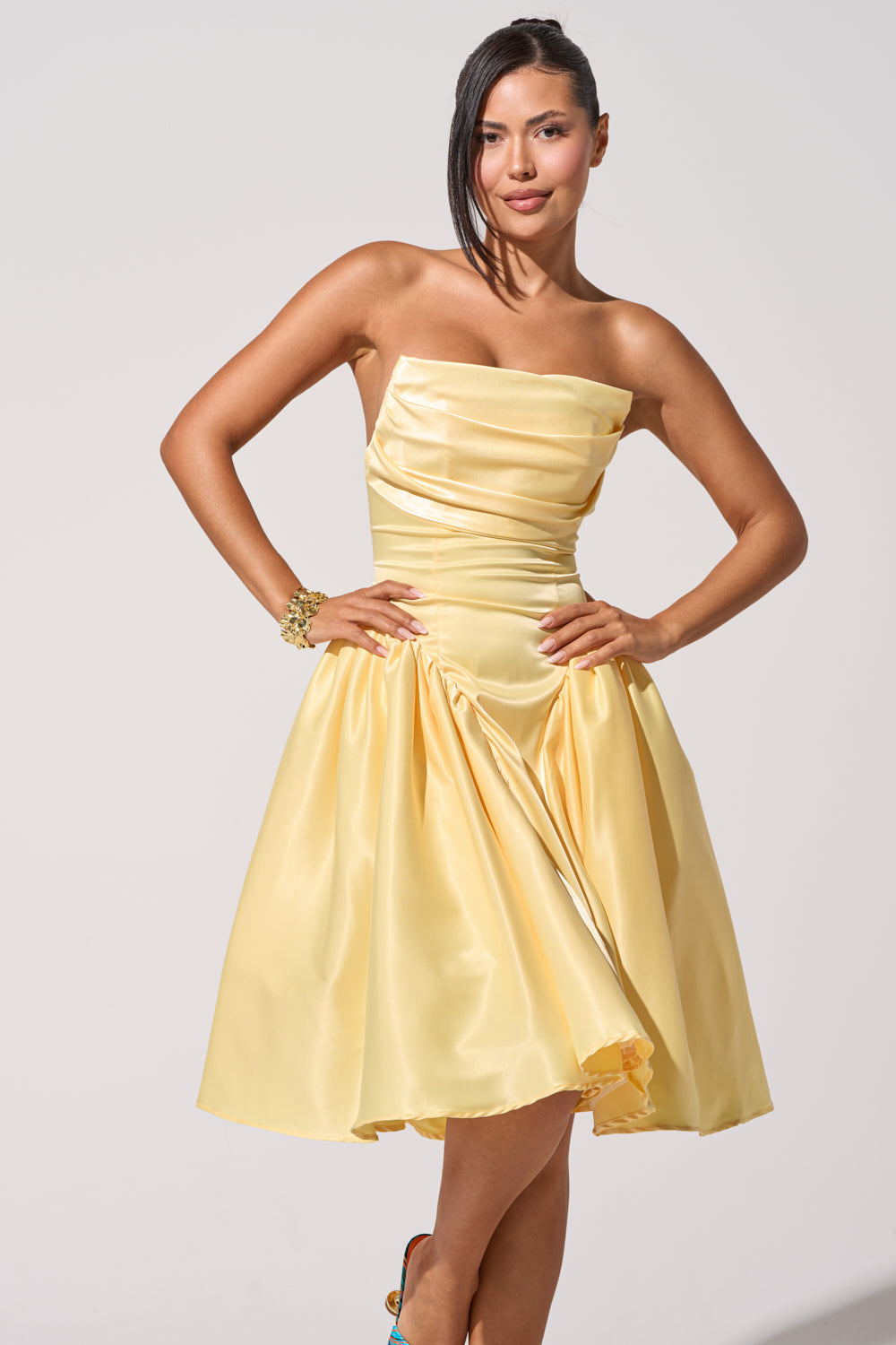 A woman poses confidently in the SHE CUTE CORSET MIDI DRESS, a yellow satin, strapless, knee-length style. With one hand on her hip and dark hair swept to the side, she stands against a plain light background.