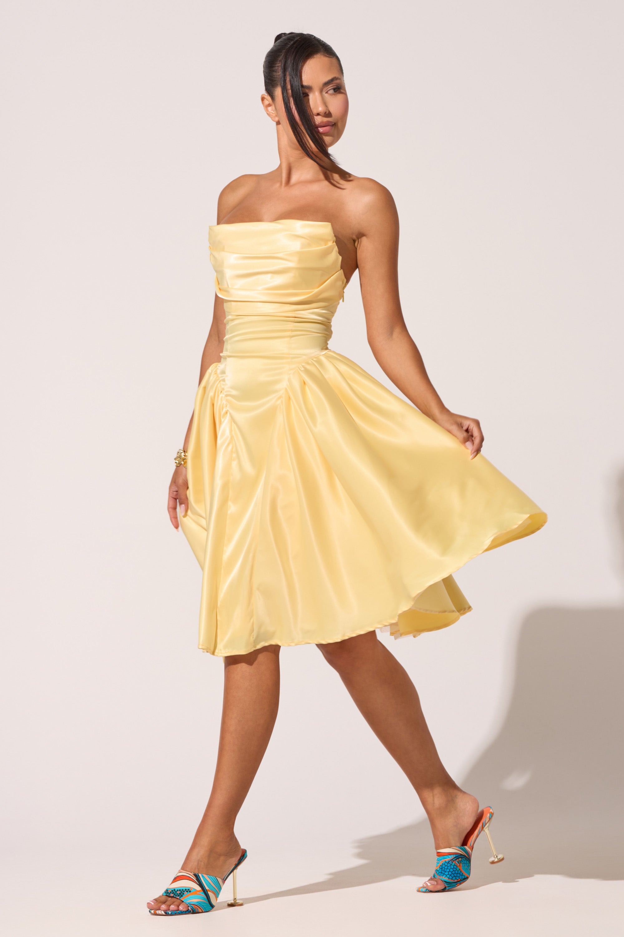 A woman poses gracefully in the SHE CUTE CORSET MIDI DRESS, a strapless yellow satin fit-and-flare style with a knee-length skirt. She pairs it with colorful high-heeled sandals against a simple light background.