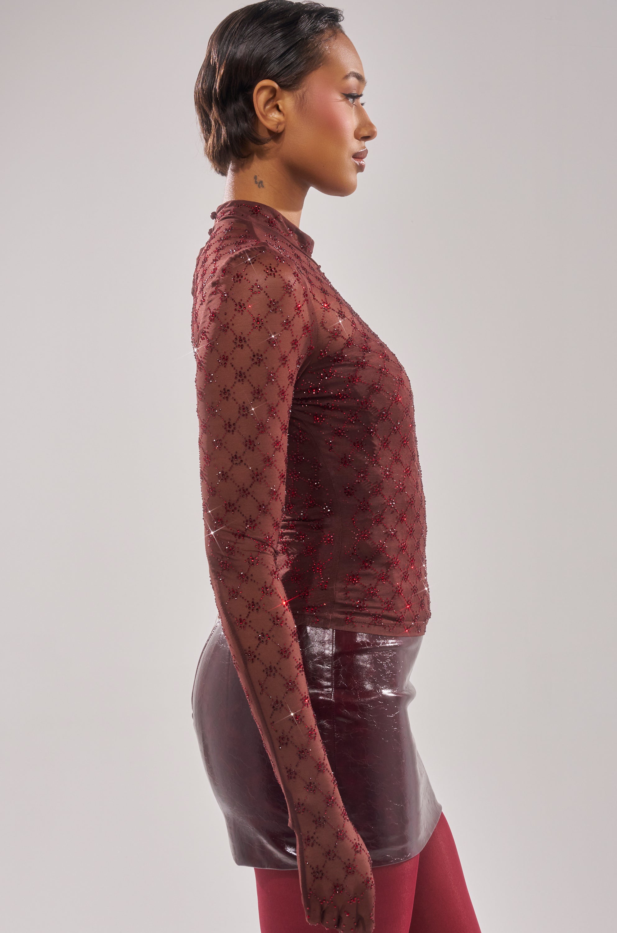A person stands in profile wearing the EXTREMELY SEXY MESH LONG SLEEVE WITH CORSET, paired with a shiny burgundy skirt and matching tights against a plain light background. 4