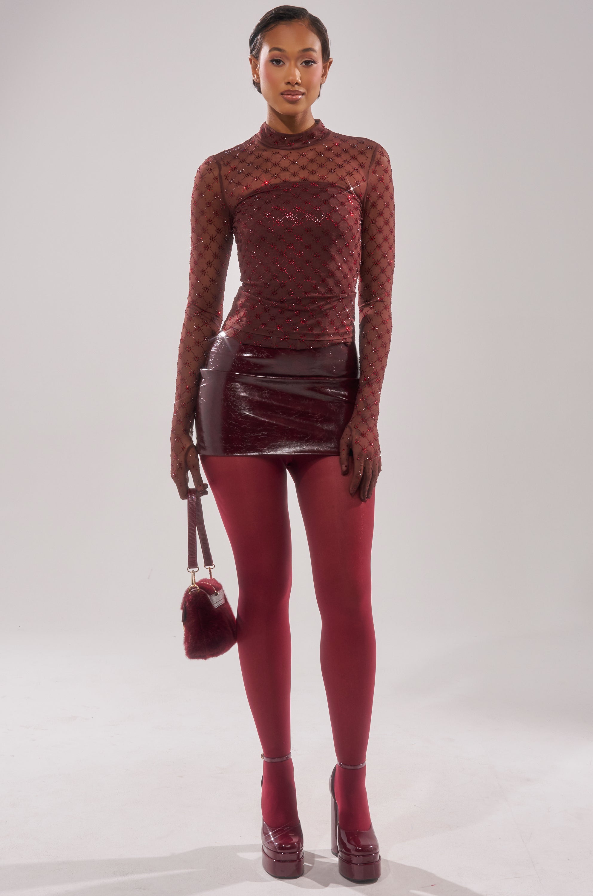 A woman in the EXTREMELY SEXY MESH LONG SLEEVE WITH CORSET, a shiny burgundy mini skirt, opaque burgundy tights, platform heels, and holding a furry burgundy purse stands against a plain white background. 6