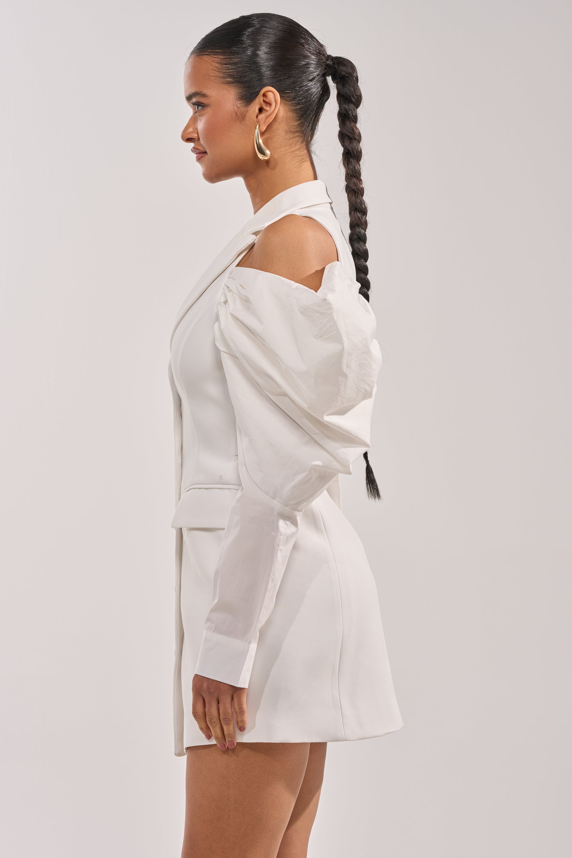 A woman in profile wears the RUNNING THINGS COLLARED MINI DRESS, a white bridal suit-inspired blazer mini with structured sleeves and a choker neckline. She has a long braid and gold hoop earrings, set against a plain background. 1