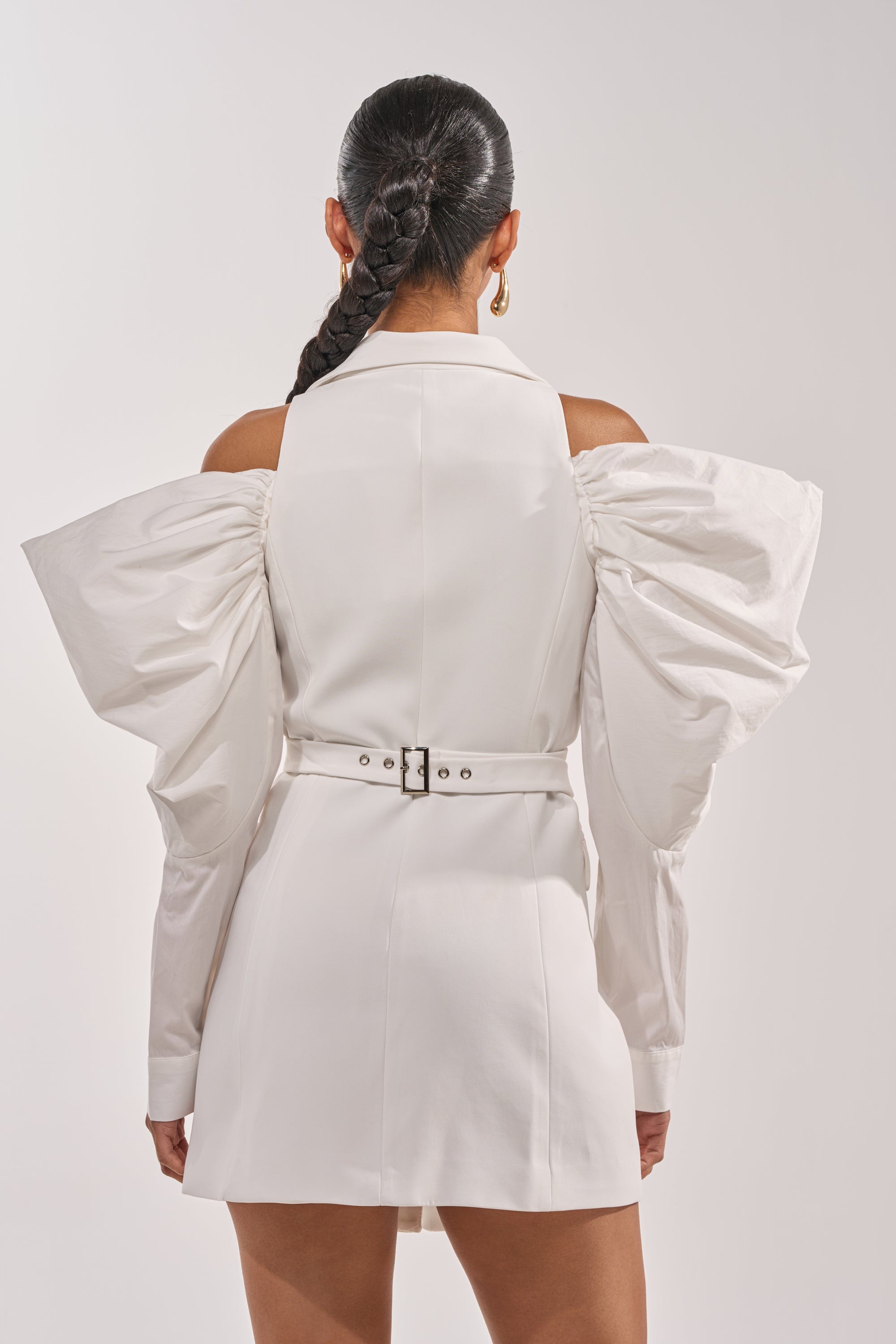 A woman with a long slicked-back braid stands facing away in the RUNNING THINGS COLLARED MINI DRESS, a white mini dress featuring dramatic structured sleeves and an open shoulder design, set against a plain light background. 2