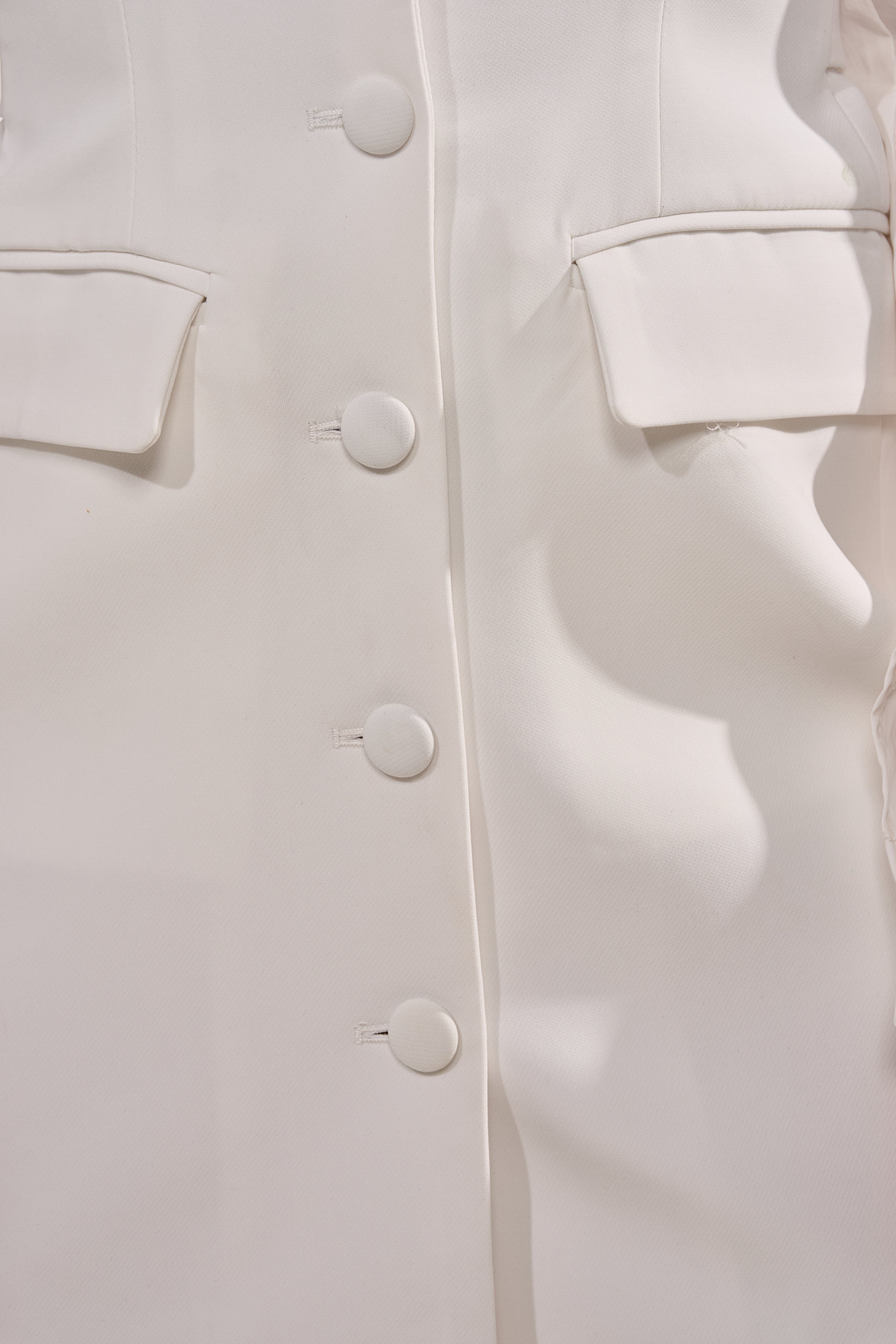 Close-up of the RUNNING THINGS COLLARED MINI DRESS, a white mini dress with five round front buttons, two flap pockets, and a smooth, structured fabric that highlights its tailored silhouette. 5