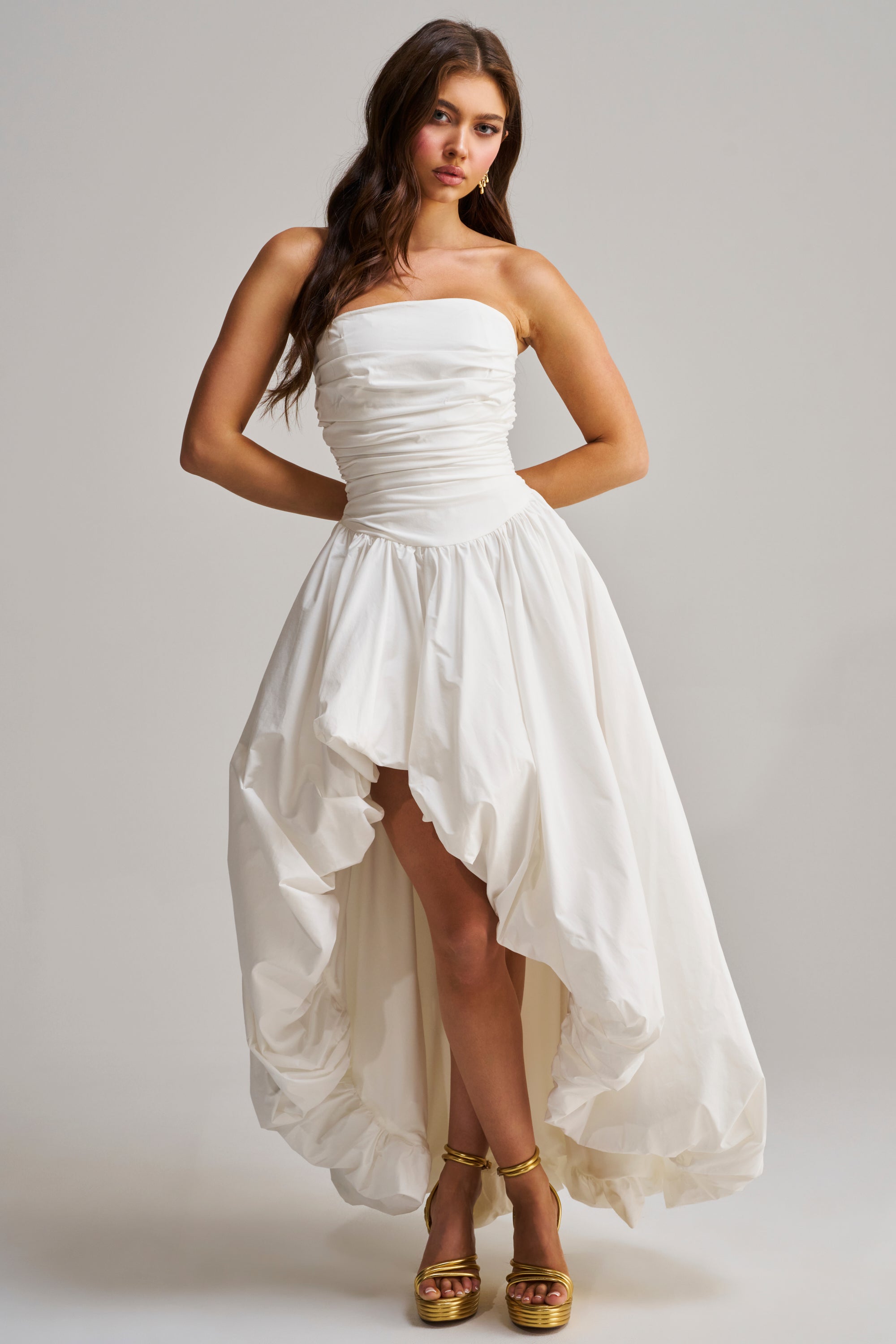 A woman with long, wavy brown hair wears the HEAD IN THE CLOUDS MAXI DRESS—a strapless white ruched maxi dress with a high-low hem and full skirt—styled with gold strappy high-heeled sandals against a plain background.