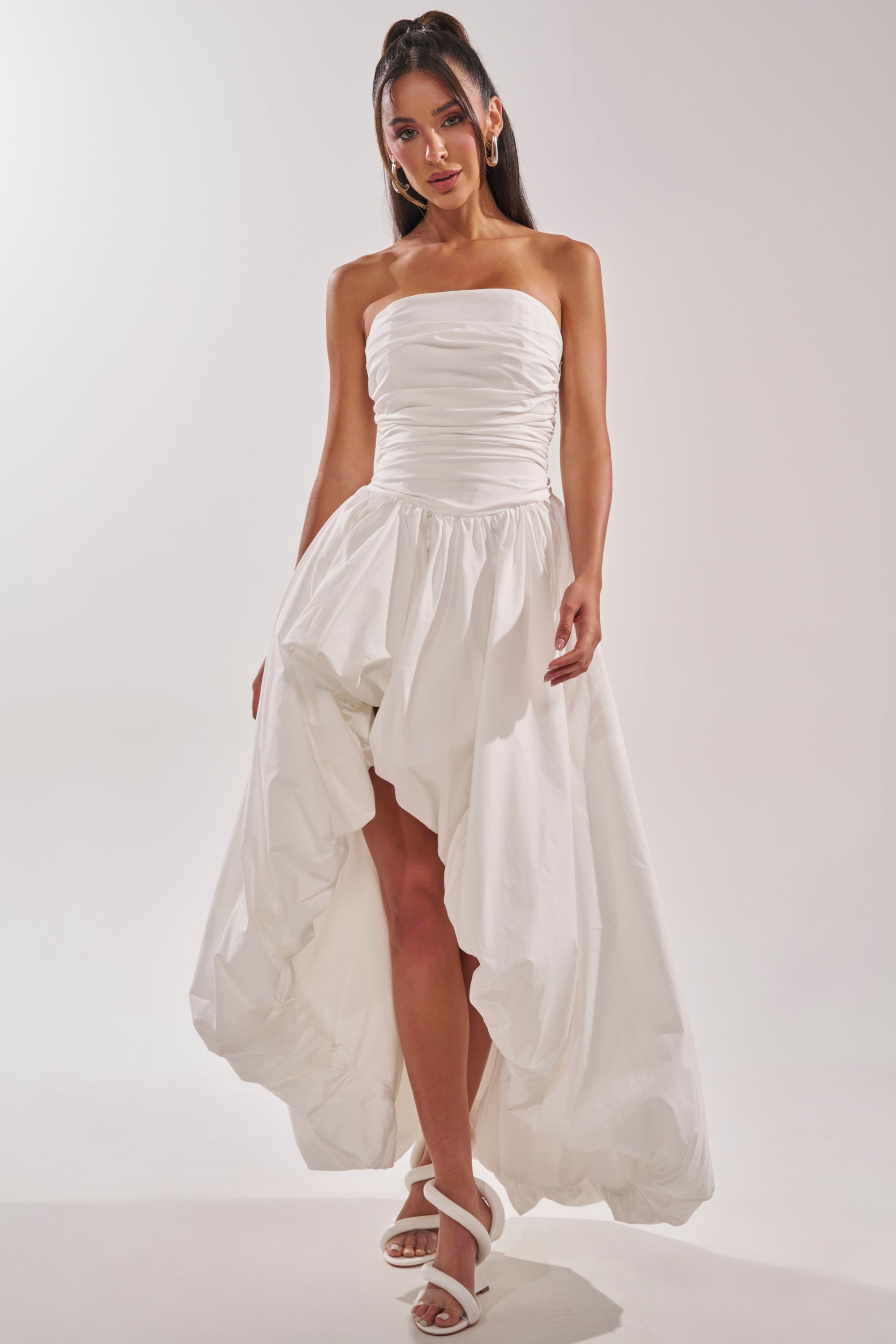 Wearing the HEAD IN THE CLOUDS MAXI DRESS, a woman styles her long dark hair up and pairs the strapless ruched maxi with earrings and white strappy heels—a chic choice for bridal formals, set against a plain background. 1