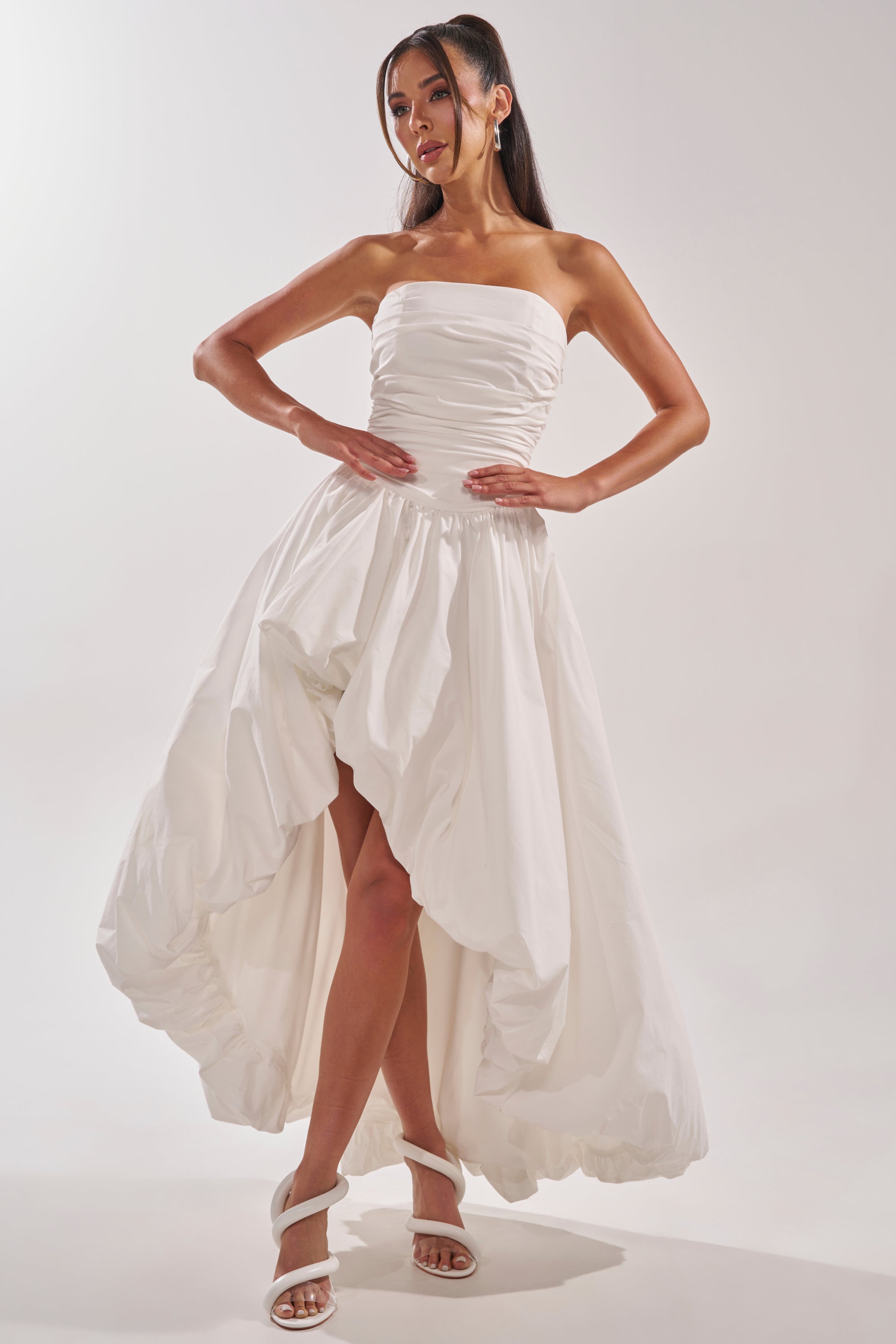 A woman models the HEAD IN THE CLOUDS MAXI DRESS, featuring a strapless ruched bodice and voluminous high-low skirt, ideal for bridal formals. She pairs it with white heeled sandals against a plain light background. 2