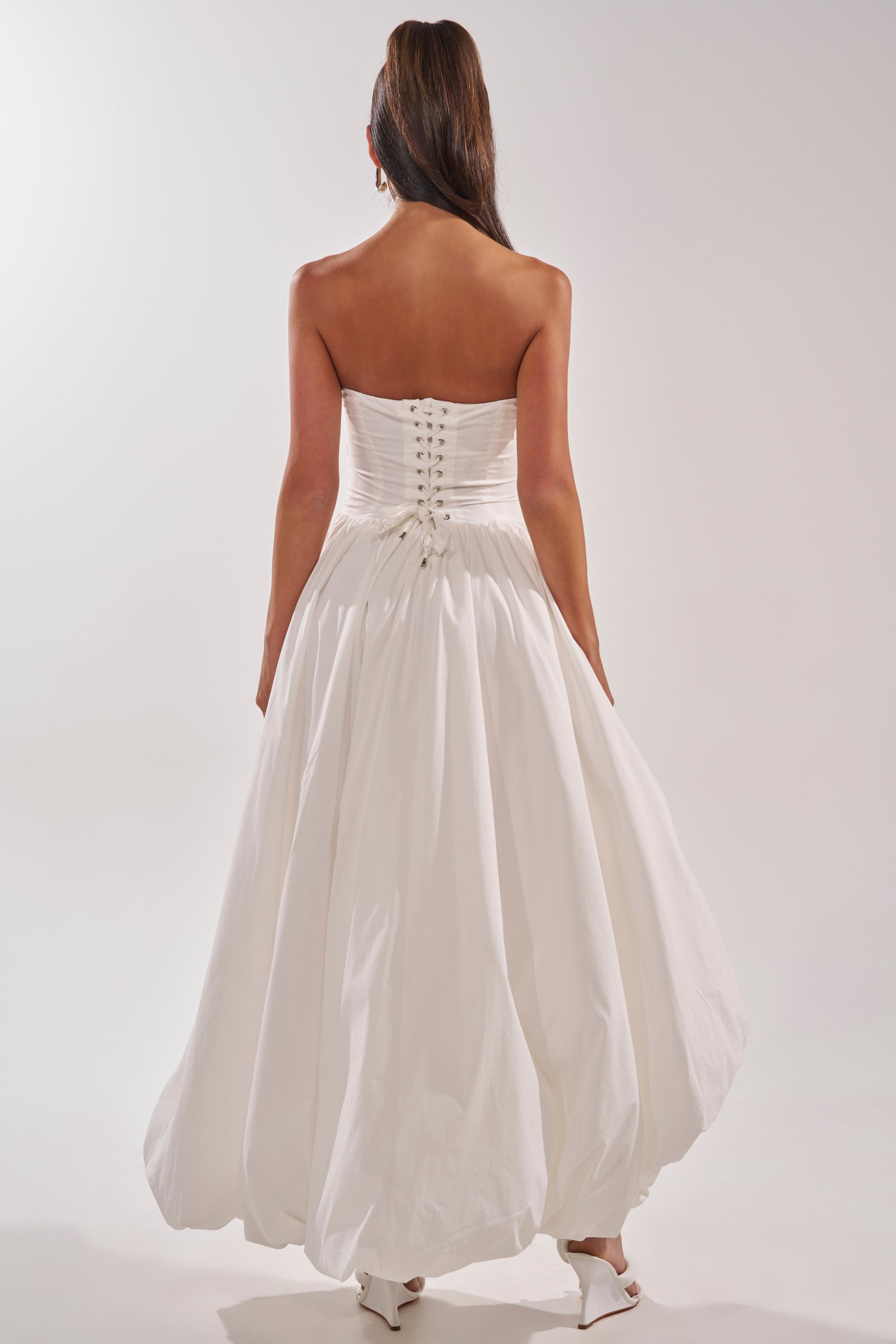 A woman with long brown hair wears the HEAD IN THE CLOUDS MAXI DRESS, showcasing its strapless corset back and full skirt as she faces away, highlighting the elegant detailing and her white heels against a plain light backdrop. 5