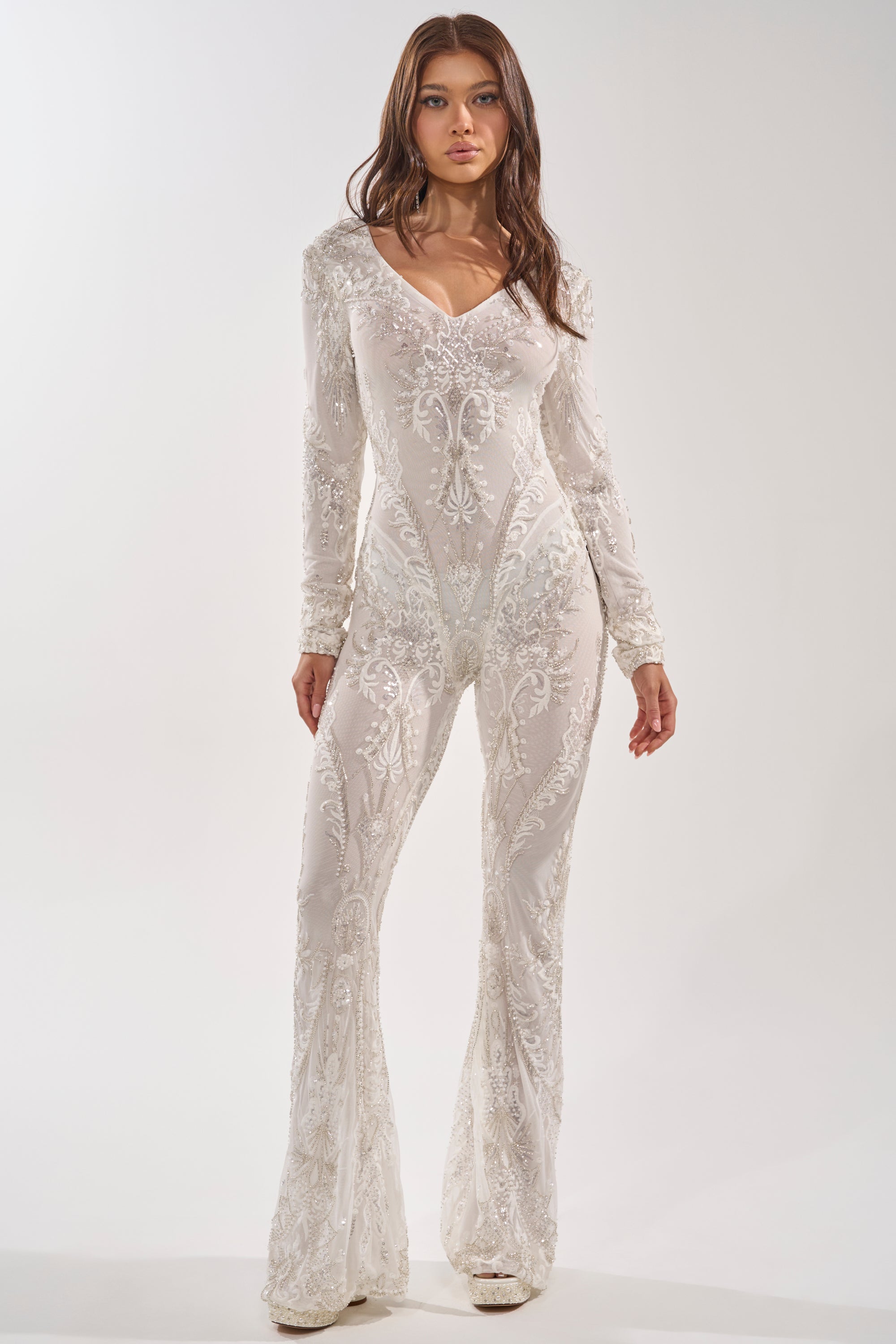 A woman poses confidently in the WATCH ME WORK EMBELLISHED SEQUIN JUMPSUIT IN WHITE, featuring a fitted silhouette, flared legs, and sparkling sequin details. She has long brown hair and neutral makeup against a plain light background. 0