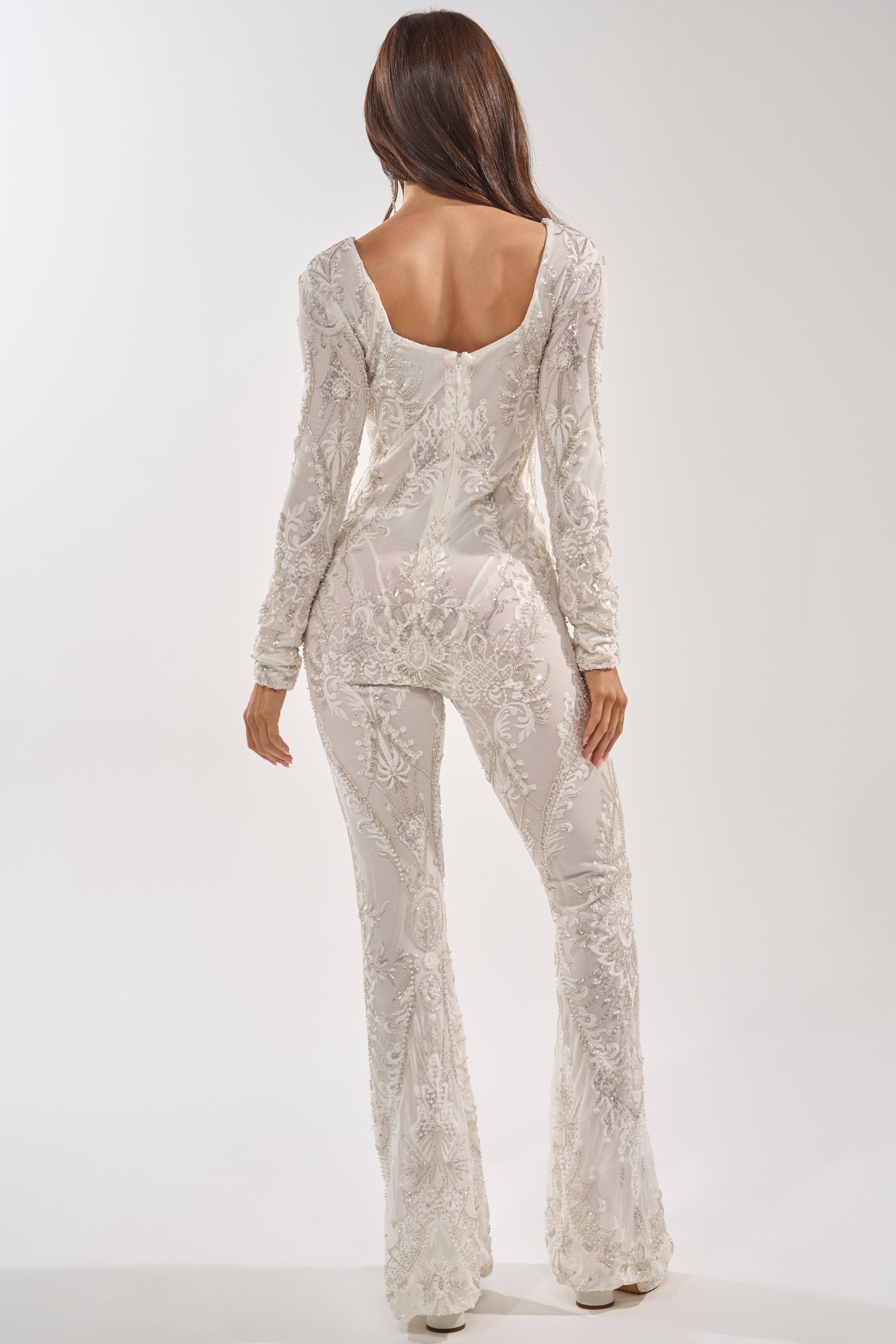 A woman with long brown hair models the WATCH ME WORK EMBELLISHED SEQUIN JUMPSUIT IN WHITE, a fitted, long-sleeve mesh jumpsuit with a low scoop back and delicate patterns, perfect for bridal formal wear, against a plain white background. 3