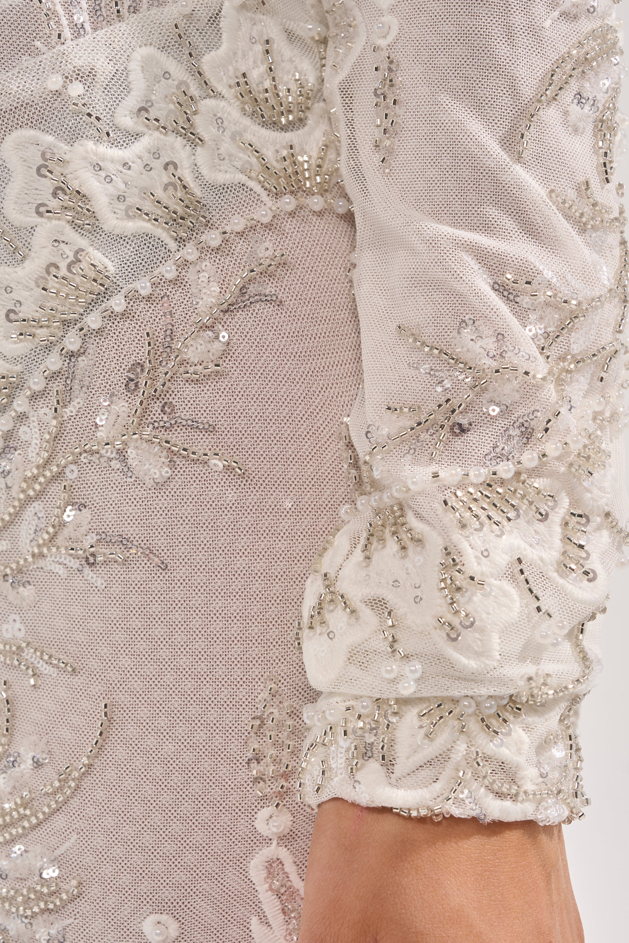 Close-up of the WATCH ME WORK EMBELLISHED SEQUIN JUMPSUIT IN WHITE, highlighting sheer white mesh with detailed floral embroidery and sequins, plus an ornate, delicate sleeve and part of the wearer’s arm. 5