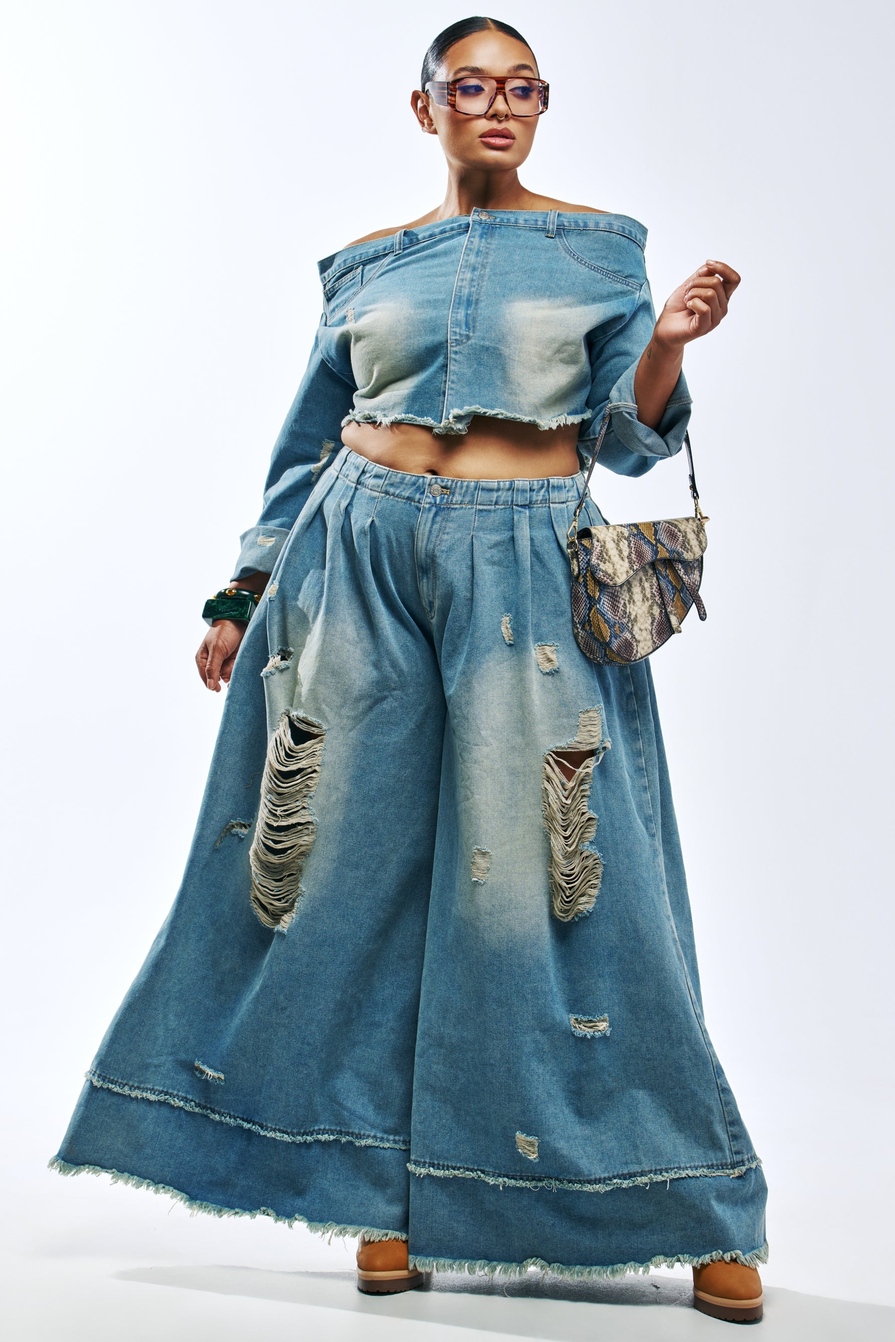 A person wearing an off-the-shoulder cropped denim jacket and CAMILLE WIDE LEG DENIM IN MEDIUM BLUE DENIM, carrying a snakeskin-print shoulder bag.