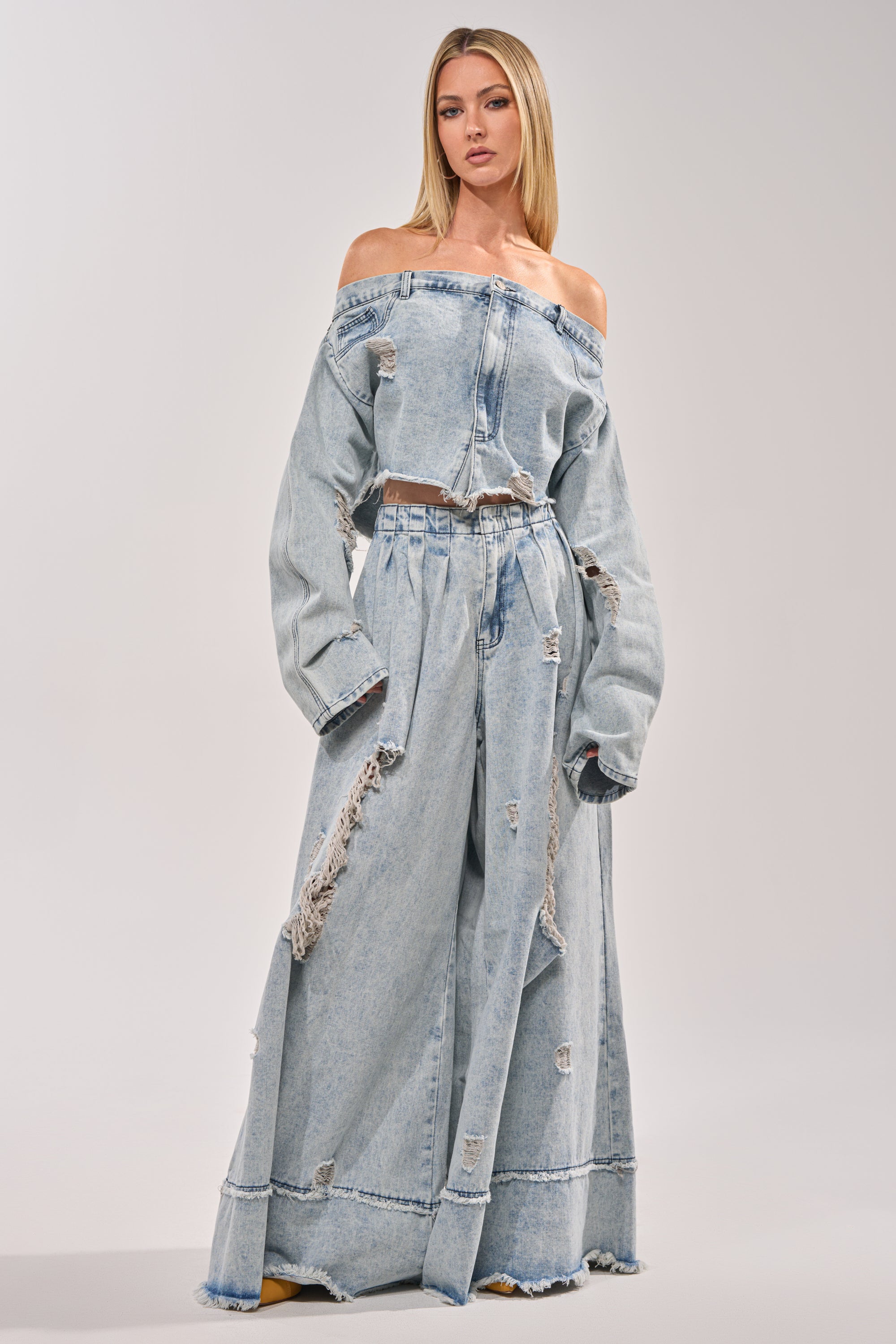 A woman stands against a plain background wearing the CAMILLE WIDE LEG DENIM—light blue, high-rise, distressed wide-leg jeans with frayed details—and an off-the-shoulder denim jacket. 0