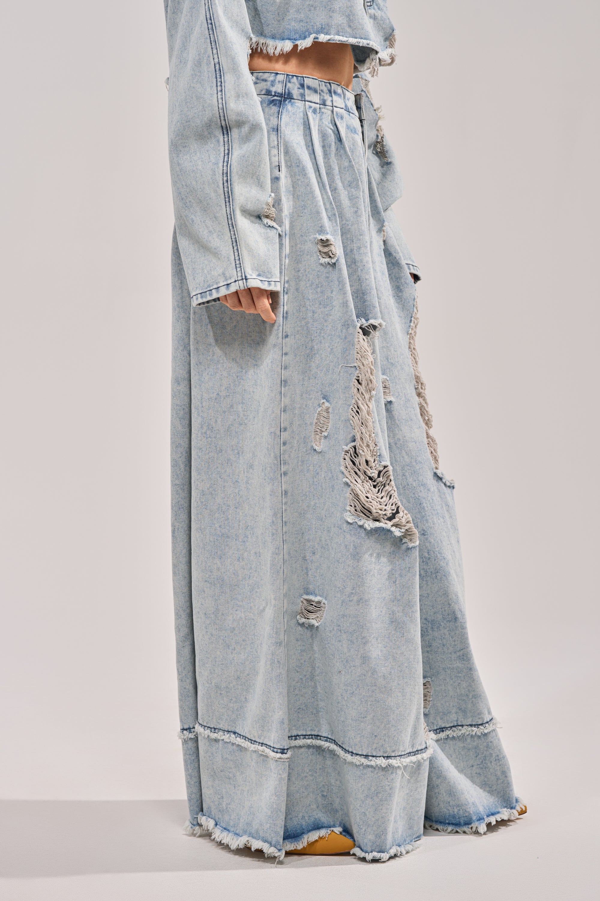 A person showcases only the lower body, wearing the CAMILLE WIDE LEG DENIM—a light blue, distressed, frayed maxi skirt style—paired with a matching long-sleeve jacket, highlighting these bold statement jeans. 2
