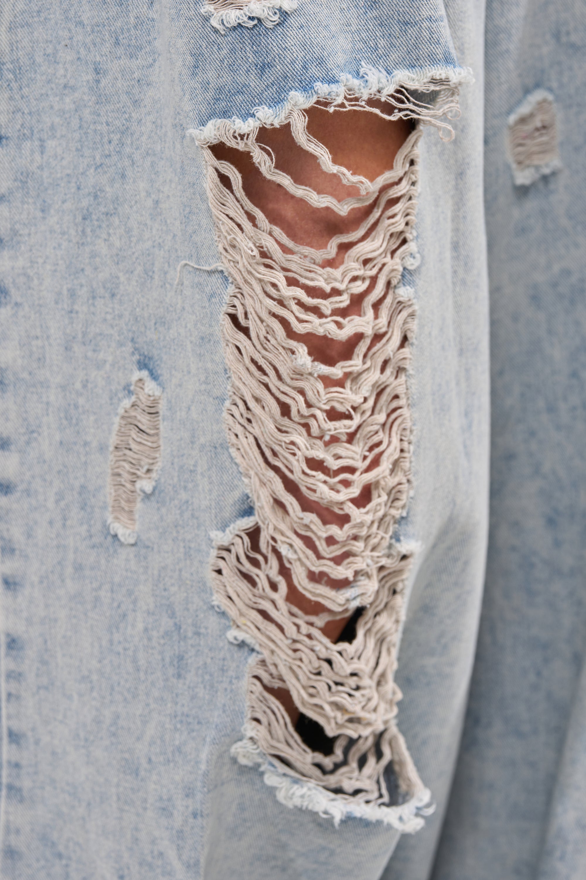 Close-up of CAMILLE WIDE LEG DENIM in light blue, highlighting the high-rise waist, wide-leg fit, a large horizontal rip showing skin, and several smaller frayed holes nearby. 6