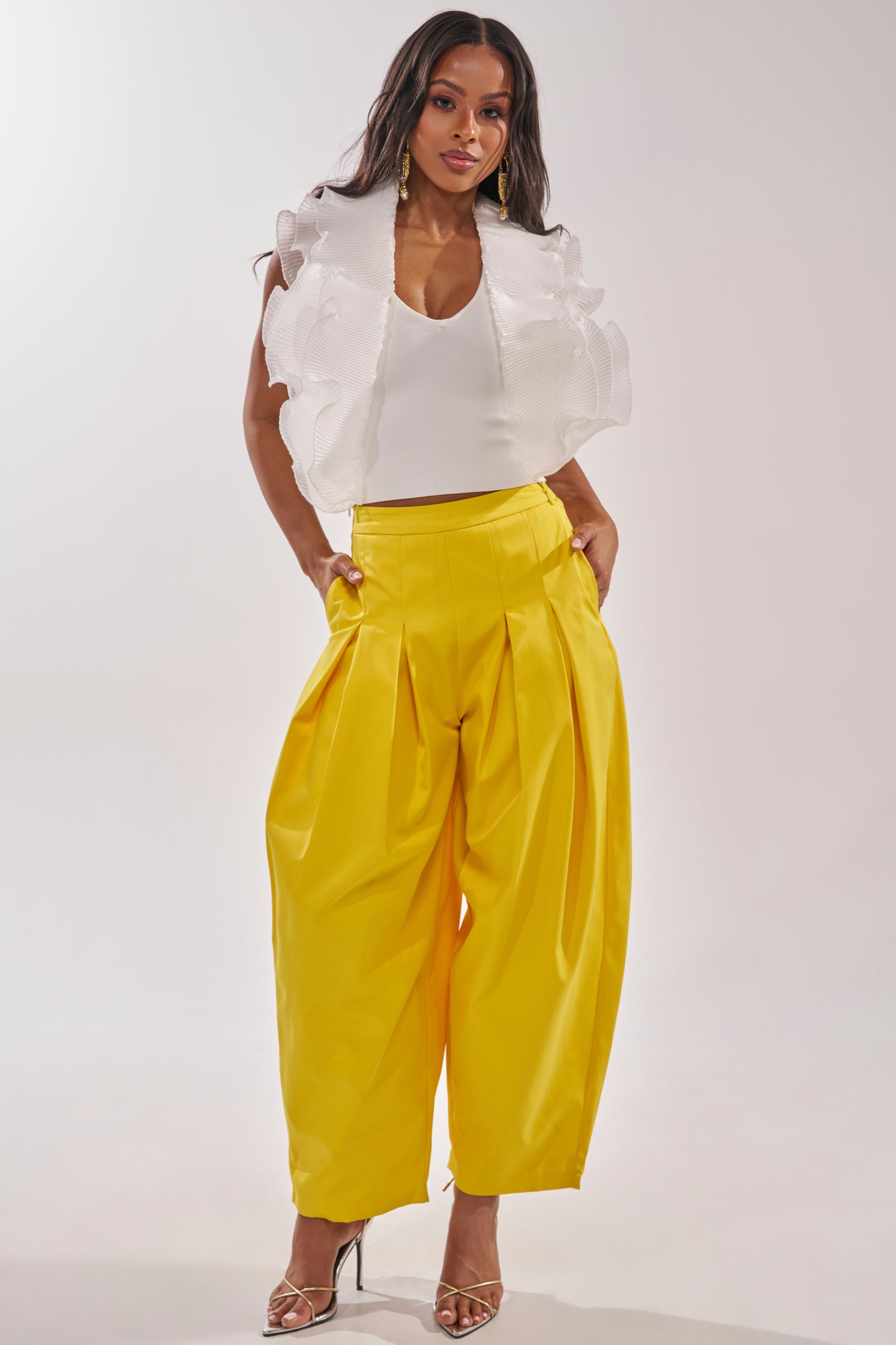 A woman stands confidently in a white sleeveless ruffled top, bright yellow BRING IT ON TROUSER with pleats, and strappy high-heeled sandals against a plain background. 0