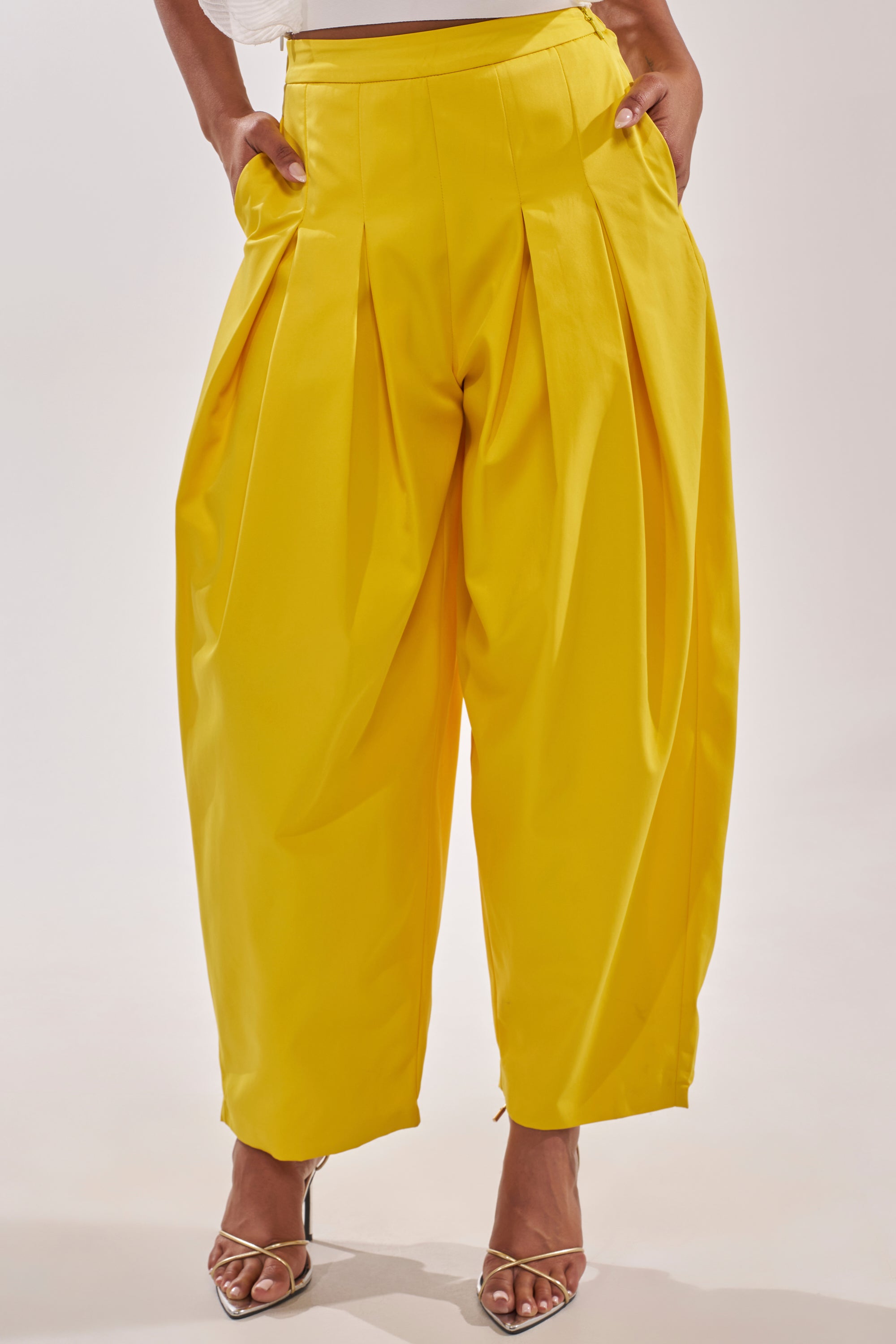 Someone wears the BRING IT ON TROUSER—bright yellow high-rise, wide-leg trousers with pleats and pockets—paired with open-toe heeled sandals. The stylish pants create a flowy silhouette; only the lower body is visible. 1