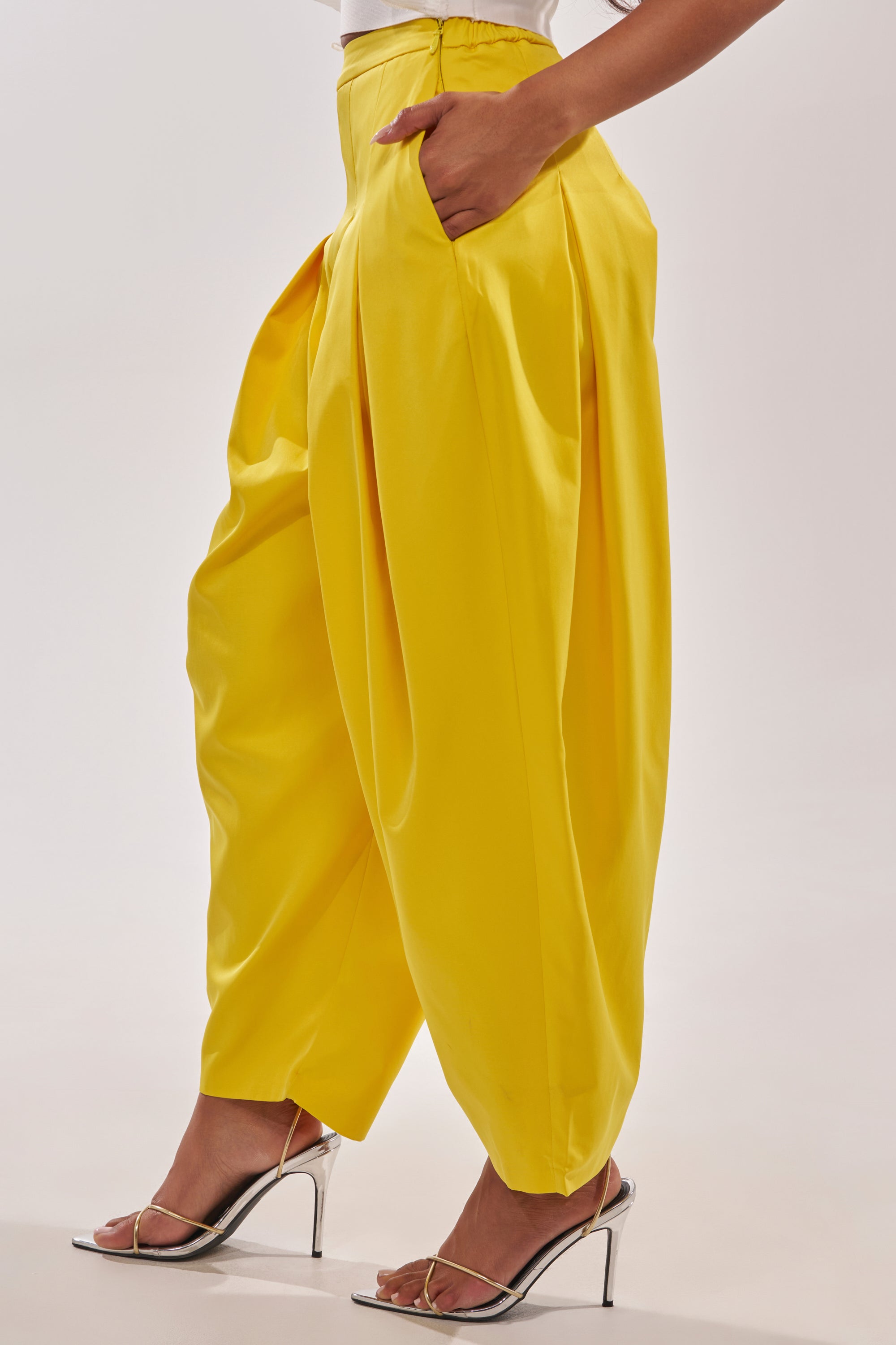 A person stands with hands in pockets, wearing the BRING IT ON TROUSER in bright yellow—high-rise, wide-leg style—paired with silver high-heeled sandals against a plain light background. 2
