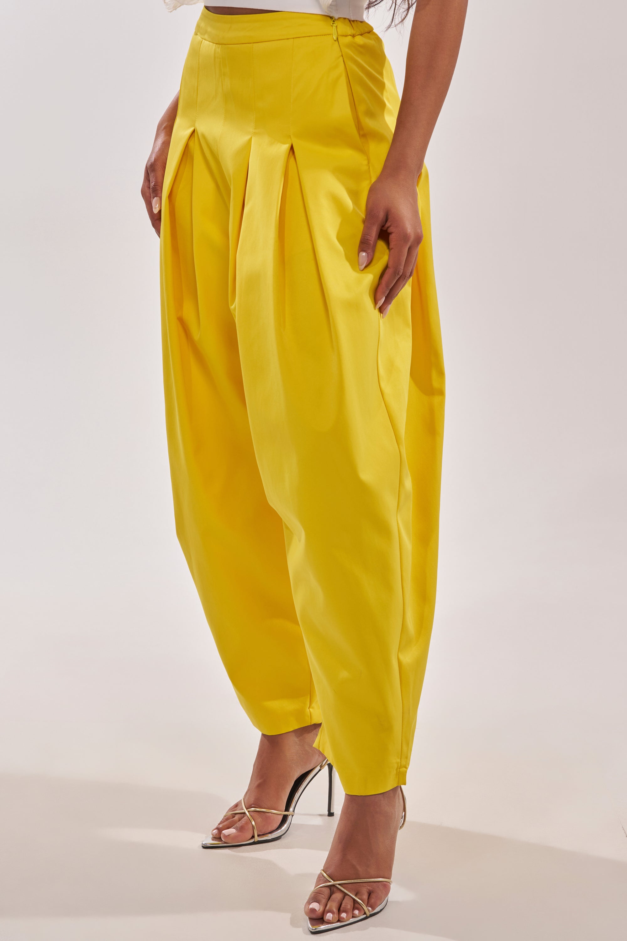 A person models the BRING IT ON TROUSER—bright yellow, wide-leg, high-rise trousers with pleats—paired with strappy silver heels. Only the lower body is shown against a plain light background. 3