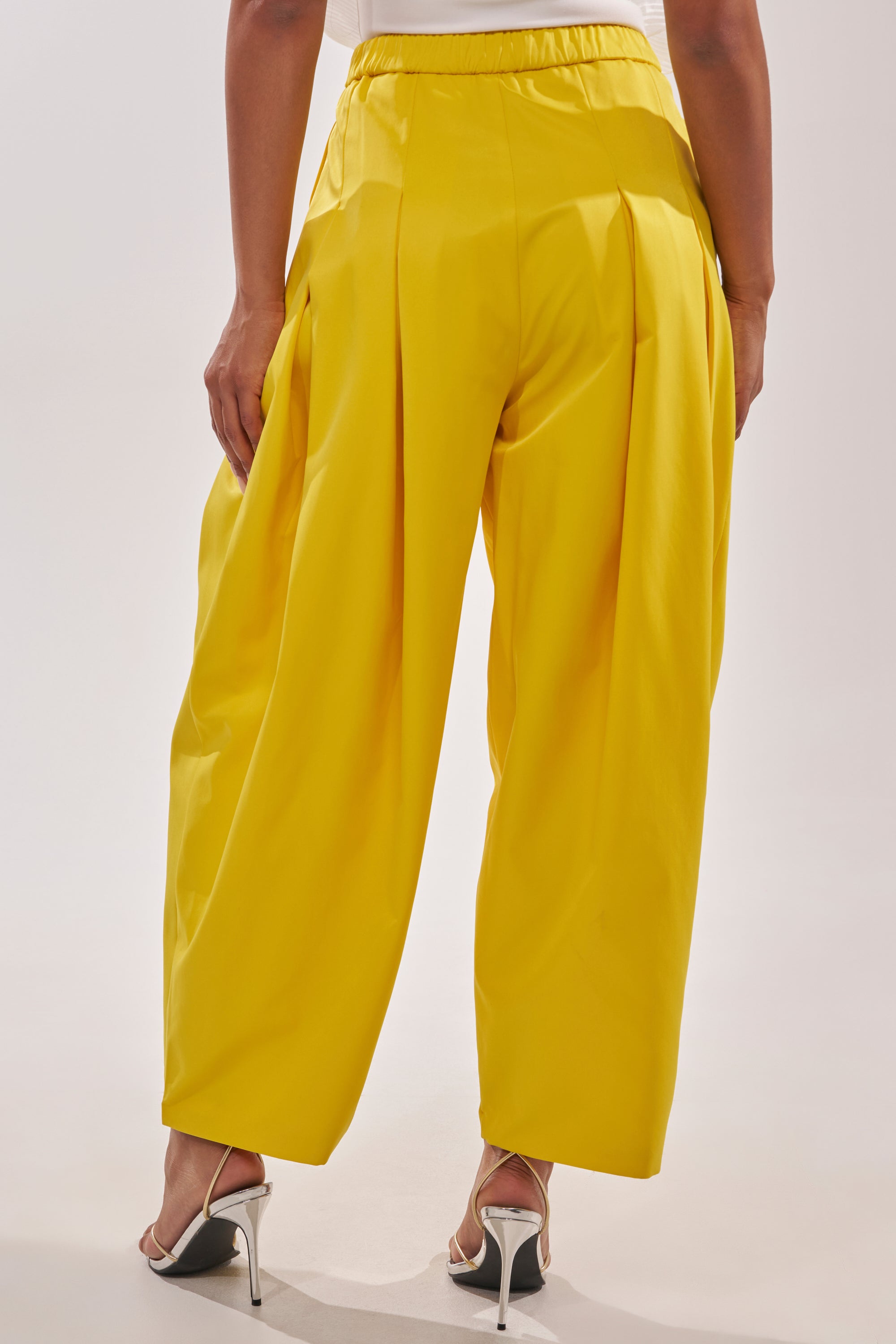 A person wears the BRING IT ON TROUSER in bright yellow with pleated details and white high-heeled sandals, standing against a plain light background. 4