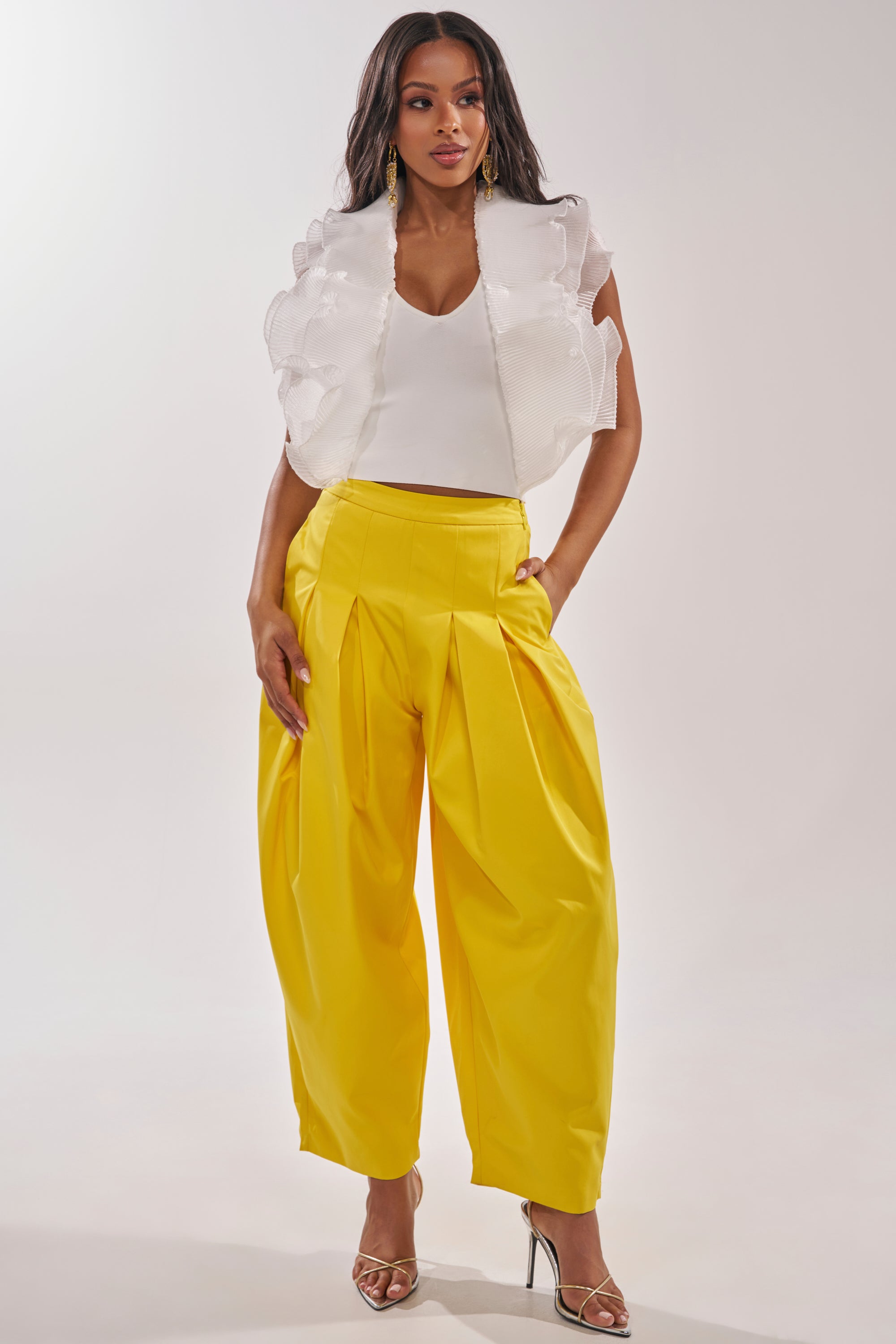 A woman models the BRING IT ON TROUSER, yellow wide-leg trousers with a high-rise fit and pleated details, styled with a white textured sleeveless top and metallic high-heeled sandals against a plain background. 5