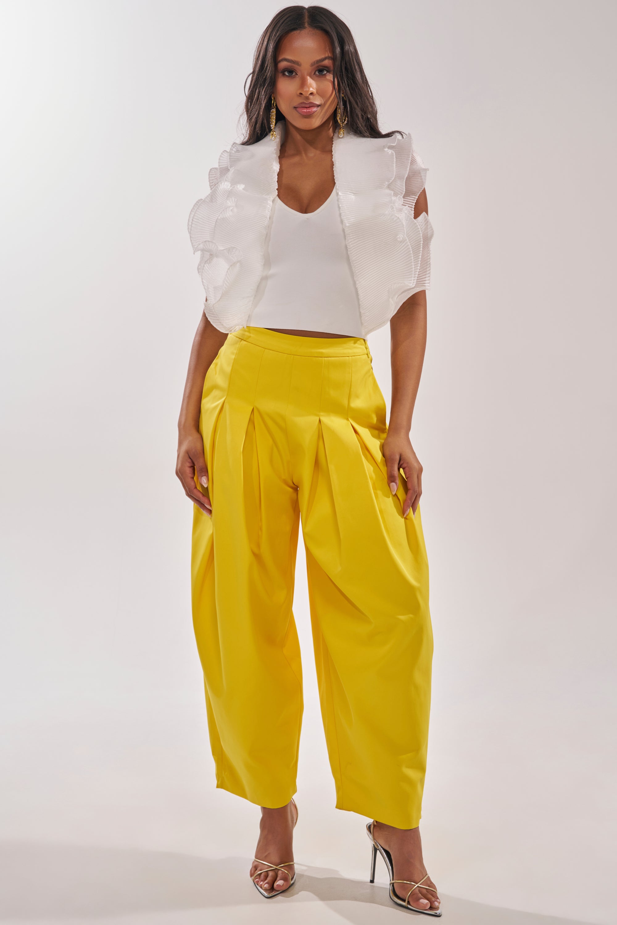A woman stands confidently in a white sleeveless top with ruffled shoulders and bright yellow BRING IT ON TROUSER featuring pleated details, paired with strappy heeled sandals against a plain background. 6