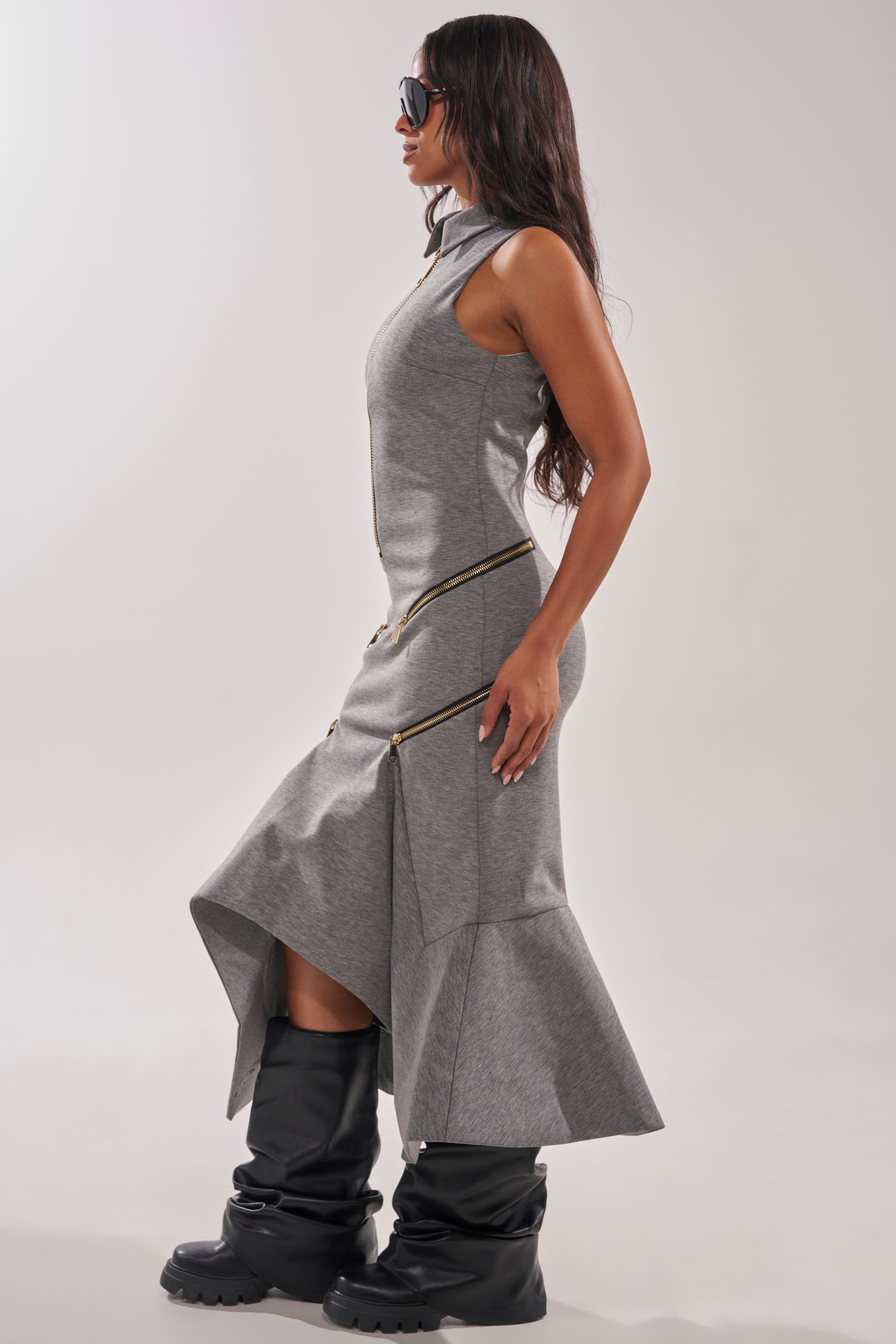 A woman in profile stands against a plain light background, wearing the ZIP CODE MAXI DRESS—a heather-gray sleeveless maxi dress with zipper details and an asymmetrical handkerchief hem—paired with black sunglasses and chunky black boots. 3