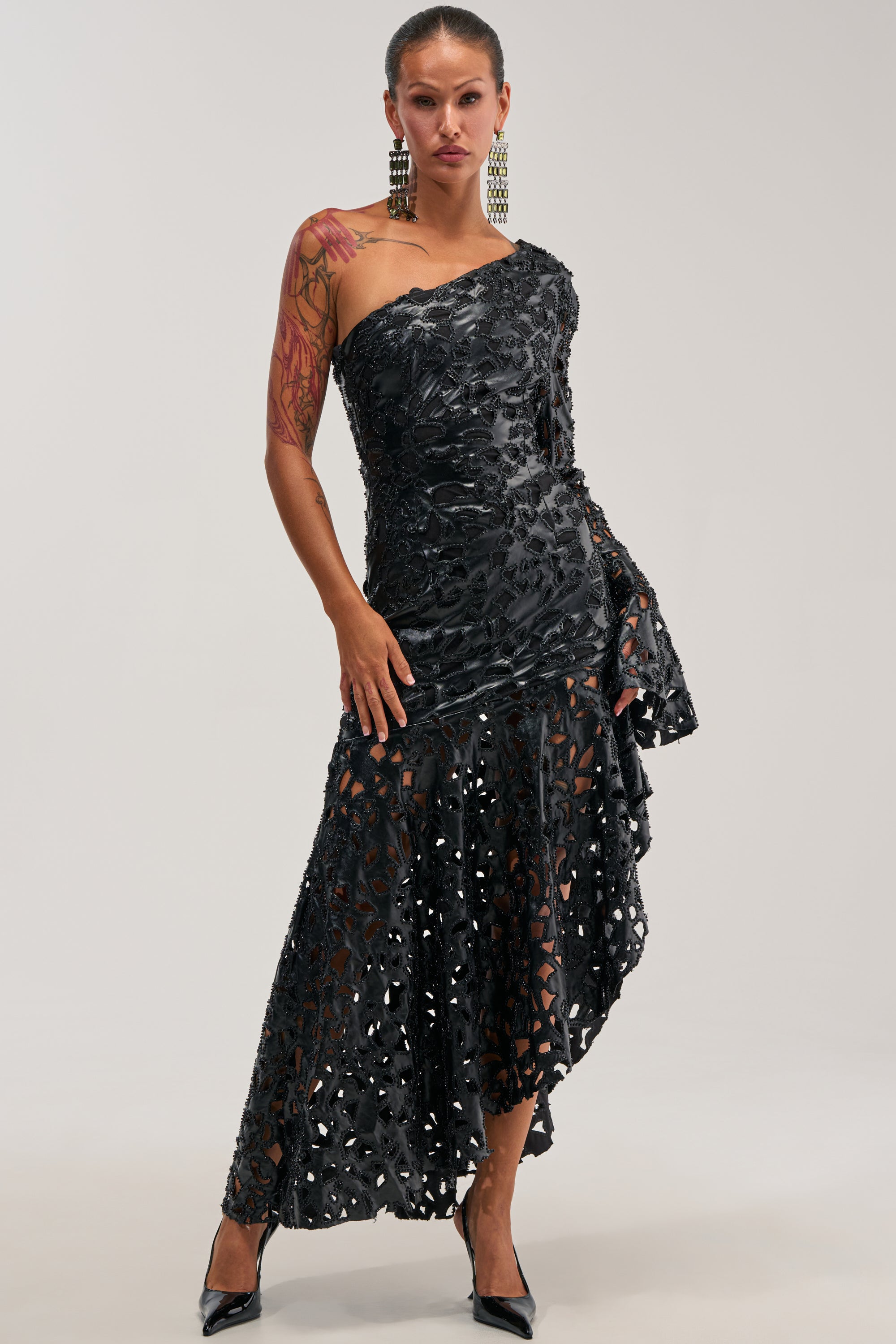 A woman poses in the MAKE A STATEMENT ONE SHOULDER MIDI DRESS, a striking black midi featuring a one-shoulder neckline, asymmetrical hem, and sheer lace panels. She accessorizes with bold rectangular earrings and black heels, while her left arm displays a red floral tattoo—making this dress an ideal choice for a winter wedding guest.