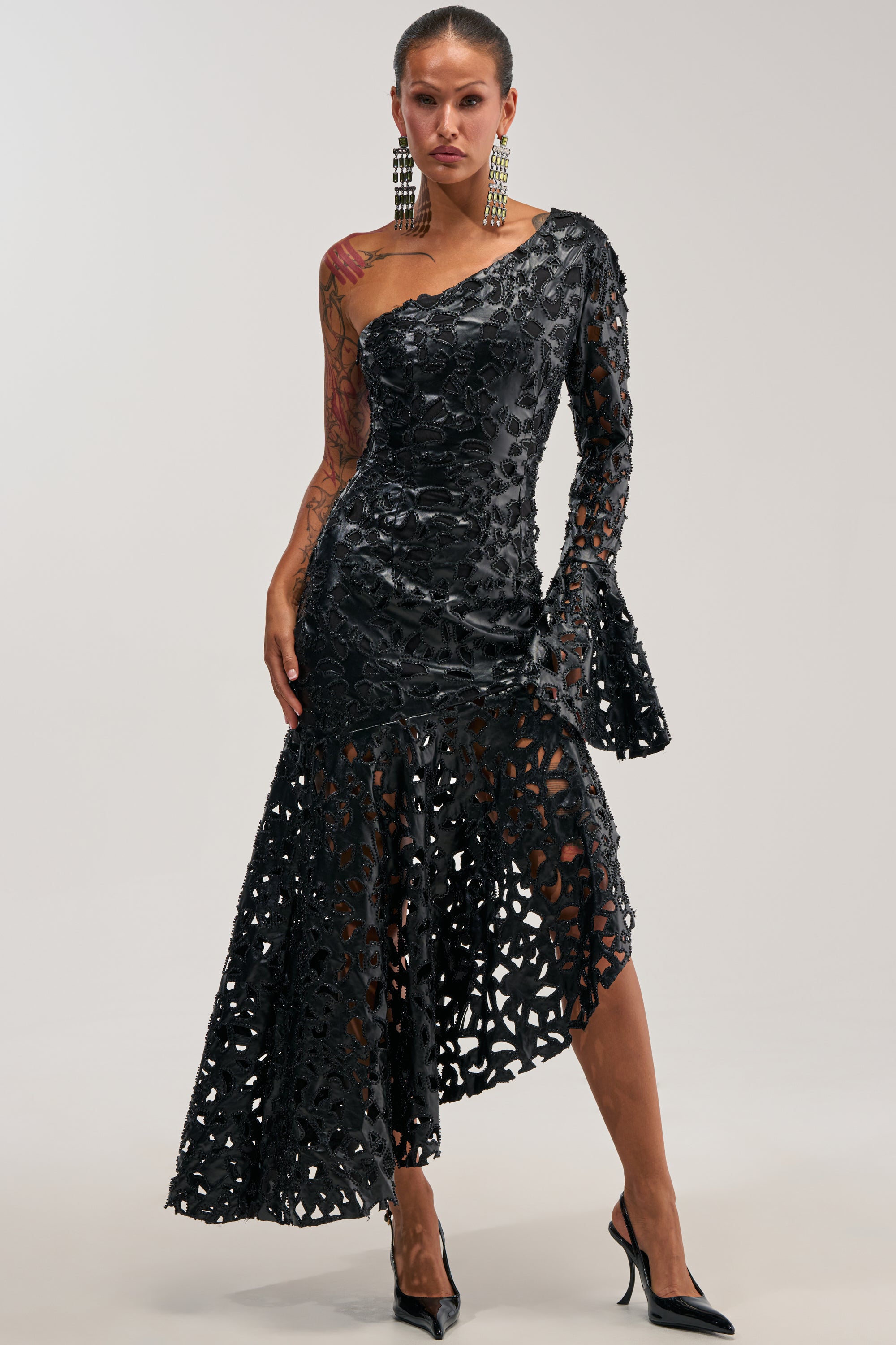 A woman poses confidently in the MAKE A STATEMENT ONE SHOULDER MIDI DRESS—a striking black, asymmetrical lace piece with a single sleeve and flared cuff. Channeling the elegance of a winter wedding guest, she completes her look with statement earrings, black heels, and visible tattoos adorning her arm, all set against a plain background.