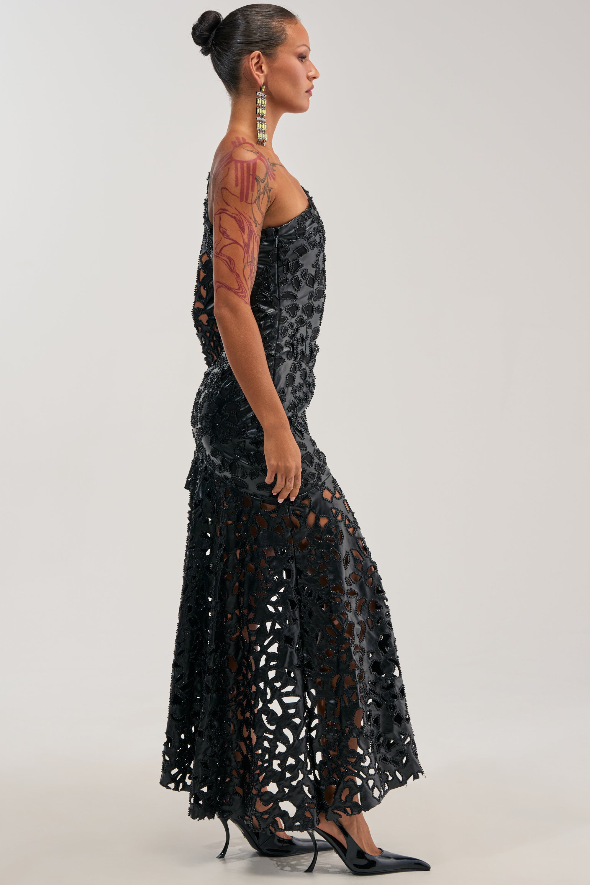 A woman with a tattooed arm, wearing the MAKE A STATEMENT ONE SHOULDER MIDI DRESS— a sleeveless, sophisticated black piece perfect for a winter wedding guest— paired with pointed heels, stands in profile against a plain background.