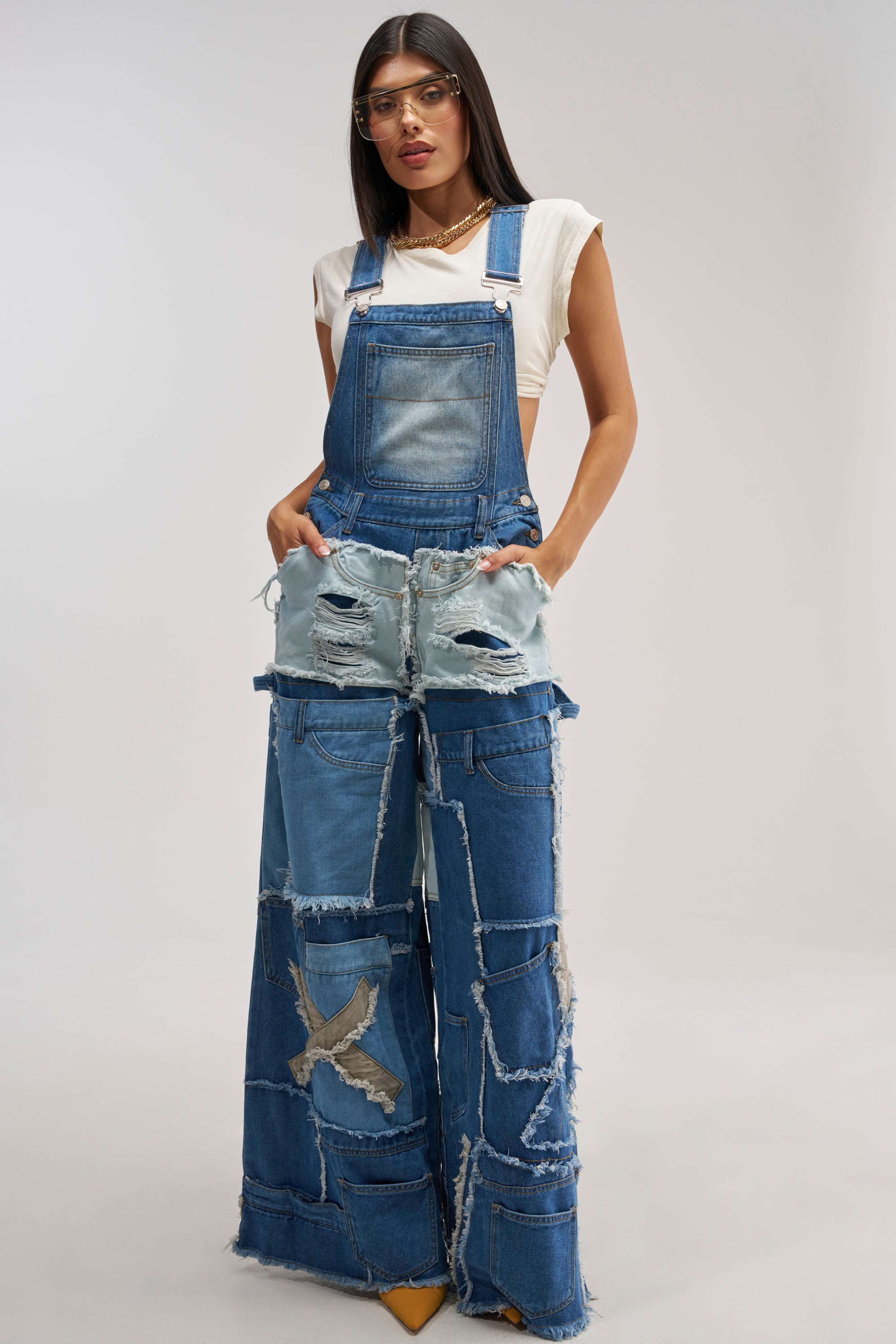 A woman with long dark hair and clear glasses stands against a light gray background, effortlessly channeling a modern streetwear vibe in the CITY CHIC DENIM OVERALLS—wide-legged patchwork denim featuring distressed details and frayed edges, paired with a white top.