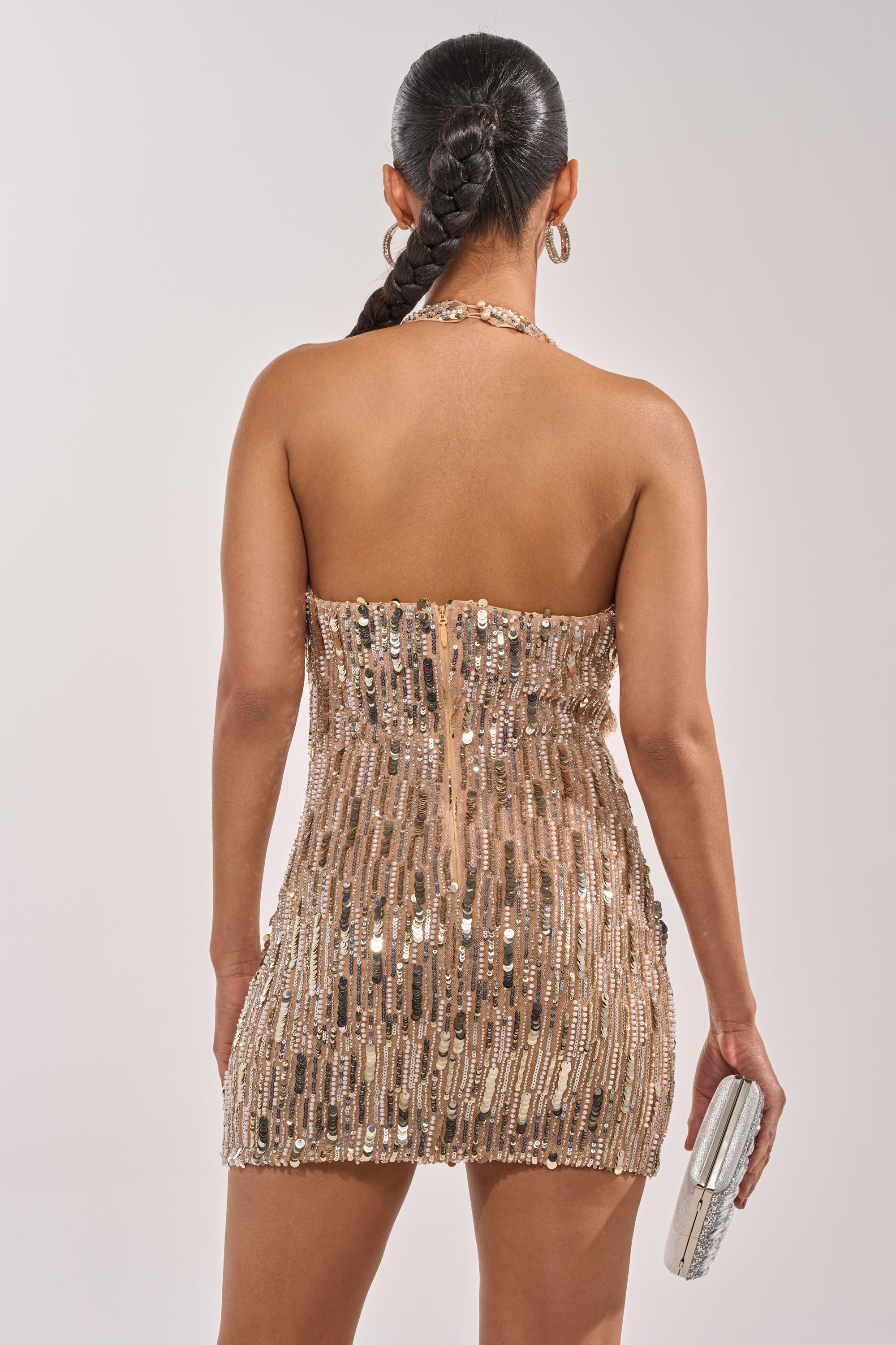 A woman with a long braided ponytail is seen from the back, wearing the AFTERPARTY SEQUIN MINI DRESS embellished in gold sequins, holding a silver clutch and hoop earrings, standing against a plain background. 3