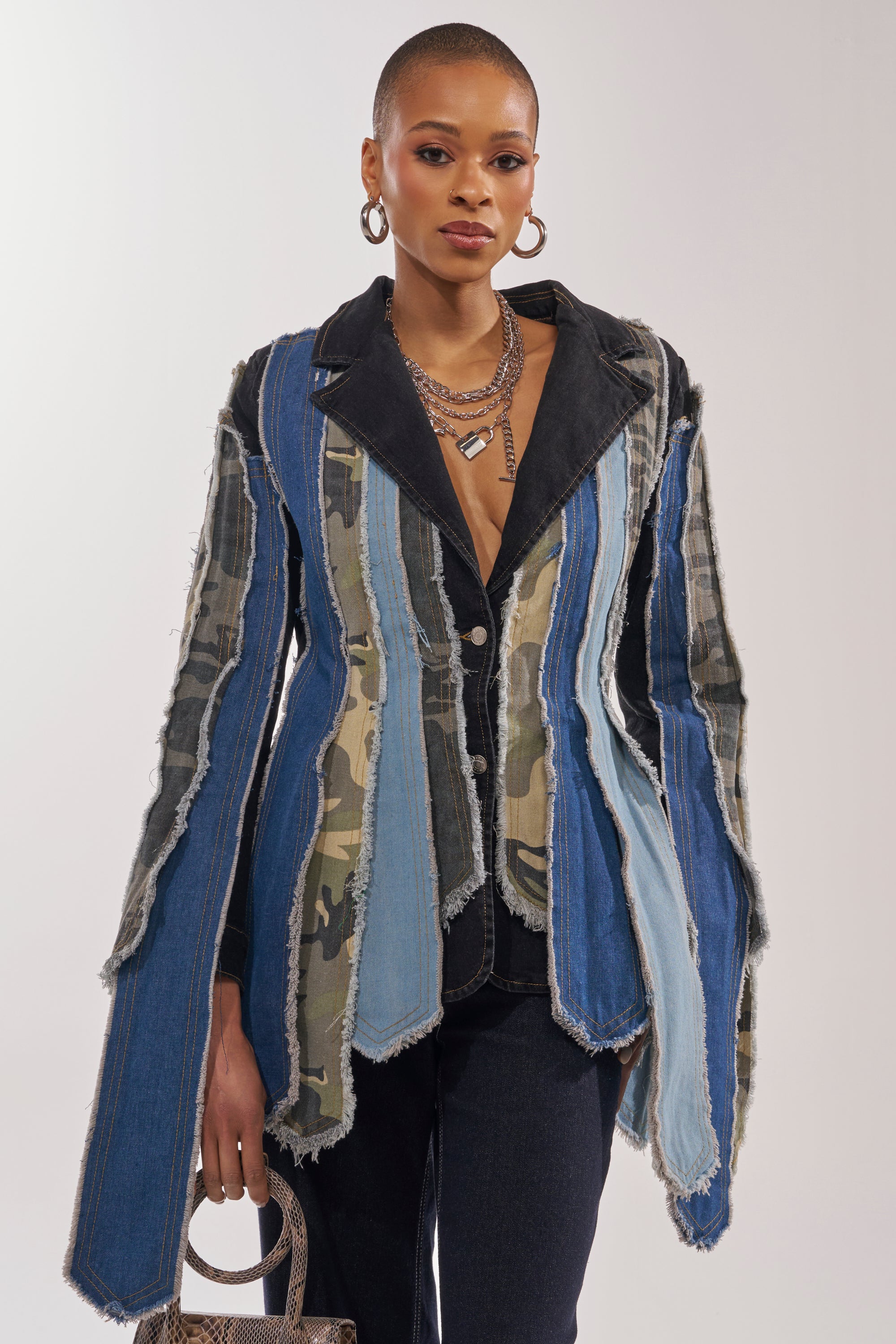 A woman with short hair models the HERE FOR IT PATCHWORK BLAZER, crafted from distressed camouflage denim, styled with dark pants, hoop earrings, layered necklaces, and a textured handbag. 0