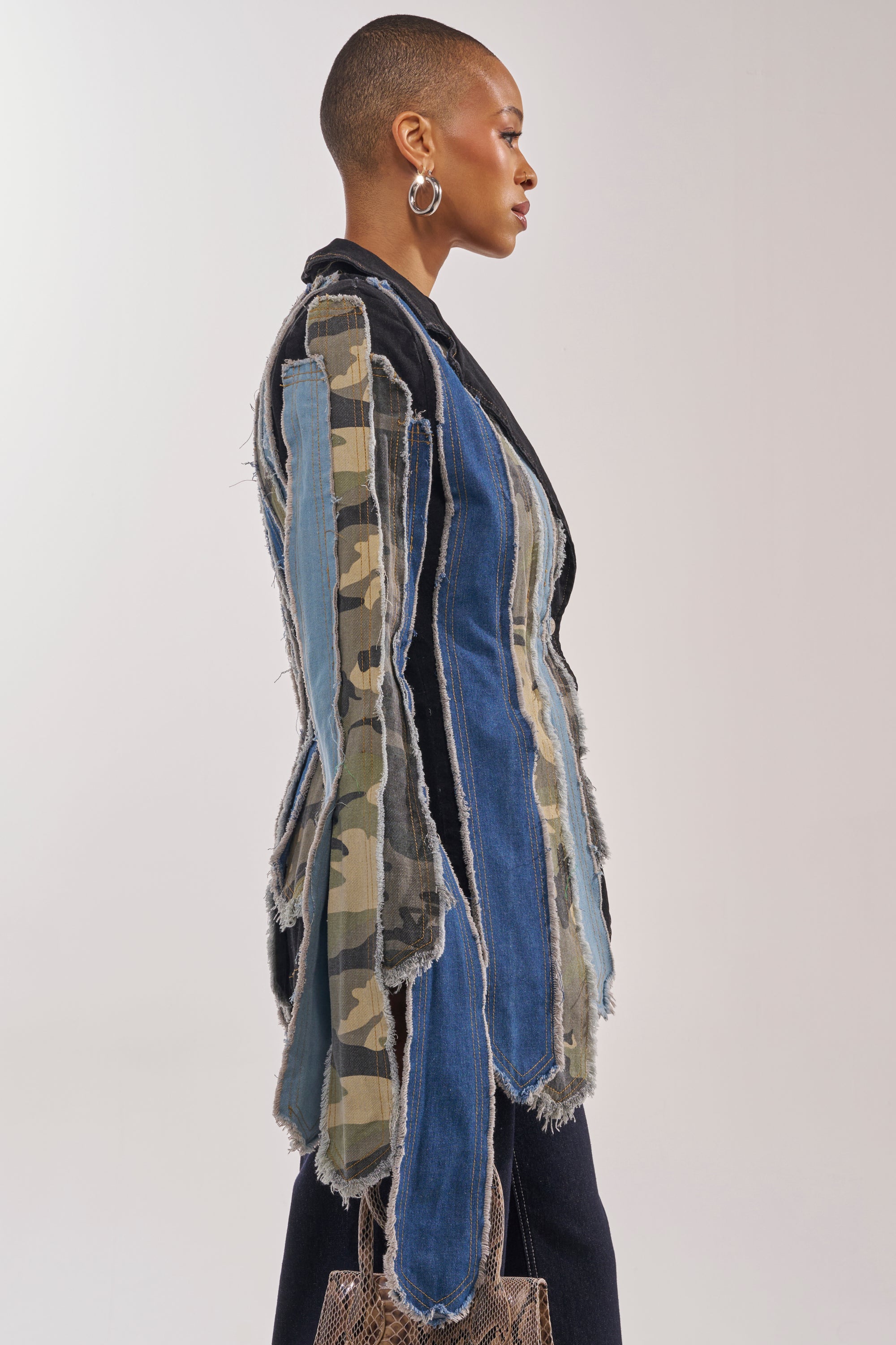 A person in profile wears the HERE FOR IT PATCHWORK BLAZER with frayed blue denim and camouflage strips, styled with short hair, large hoop earrings, and a small textured bag against a plain background. 1